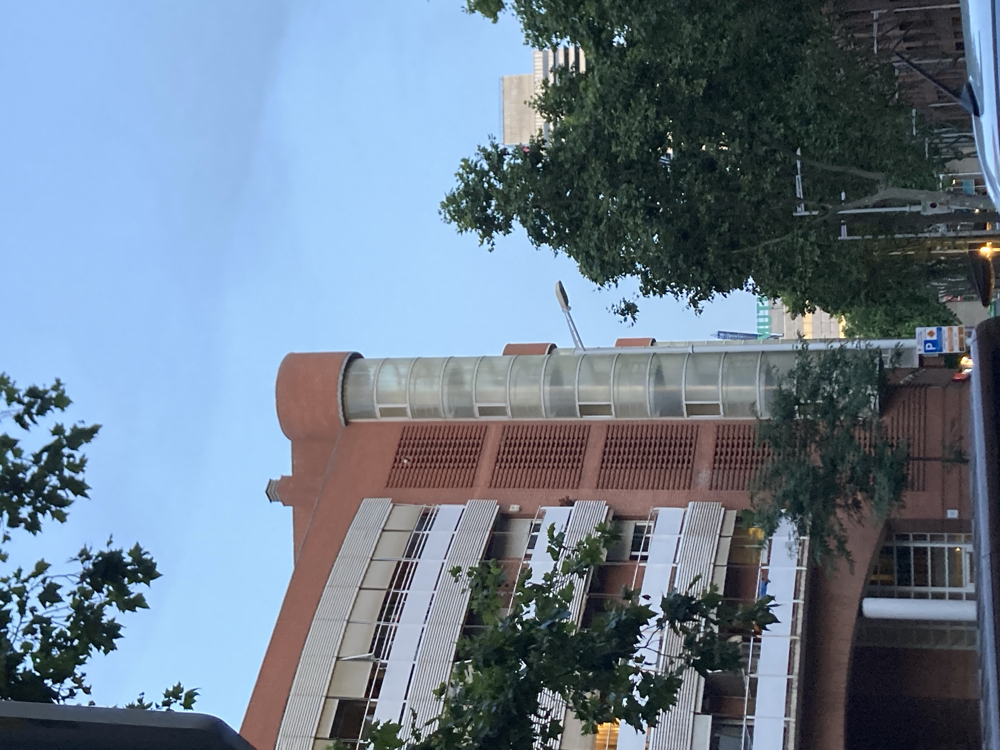The image captures a street view in Barcelona, Spain, under a slightly overcast sky. The foreground is dominated by the side of a reddish-brown brick building with multiple floors featuring white balconies and large windows. Adjacent to this building is a curved glass structure, possibly a staircase or elevator shaft, with multiple panes. Trees with lush green foliage are prominent, partially obscuring the building and other structures in the background. In the distance, another taller building is visible, with a sign that appears to read "MUTUAL". Streetlights and traffic signs are also present, indicating an urban environment. The scene suggests a daytime setting, likely late afternoon or early evening, given the lighting conditions. There are no visible people or specific actions taking place.