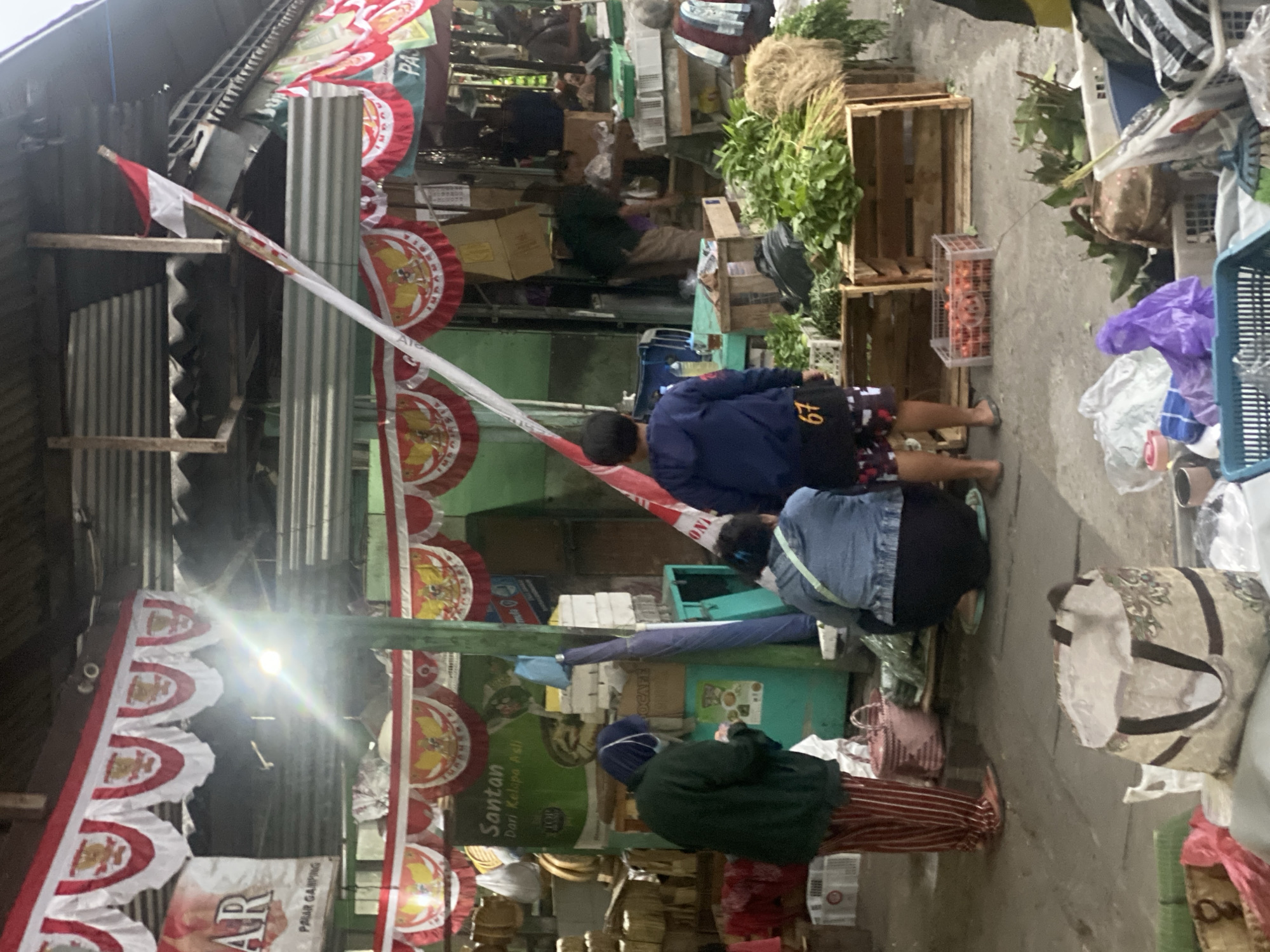 The image captures a scene at a traditional market in Ambarket-awang, Indonesia, likely during a festive occasion, given the prominent Indonesian flags and red and white bunting. The market appears to be an open-air covered structure with a corrugated metal roof supported by wooden beams and metal poles.
In the foreground, a large, patterned tote bag sits open on the ground, partially filled with items. To its left, a red plastic bag and a woven basket are visible.
The central area of the image focuses on several individuals interacting around market stalls. On the left, a person wearing a dark green jacket, a headscarf, and striped trousers is looking down at something in their hands. They are standing next to a pink woven basket. Beside them, another person, wearing a blue denim jacket and black trousers, is partially obscured, appearing to be seated or kneeling. In the center, a young person in a blue hooded jacket with the number "67" on their shorts stands facing away from the camera, observing the market activities.
Market stalls are laden with goods. Wooden crates are stacked, some containing produce like tomatoes and leafy greens. Further back, other vendors and shoppers are visible, some attending to their wares, and others browsing. The overall atmosphere suggests a busy marketplace environment. The presence of Indonesian flags and decorative bunting suggests a national holiday or celebration.