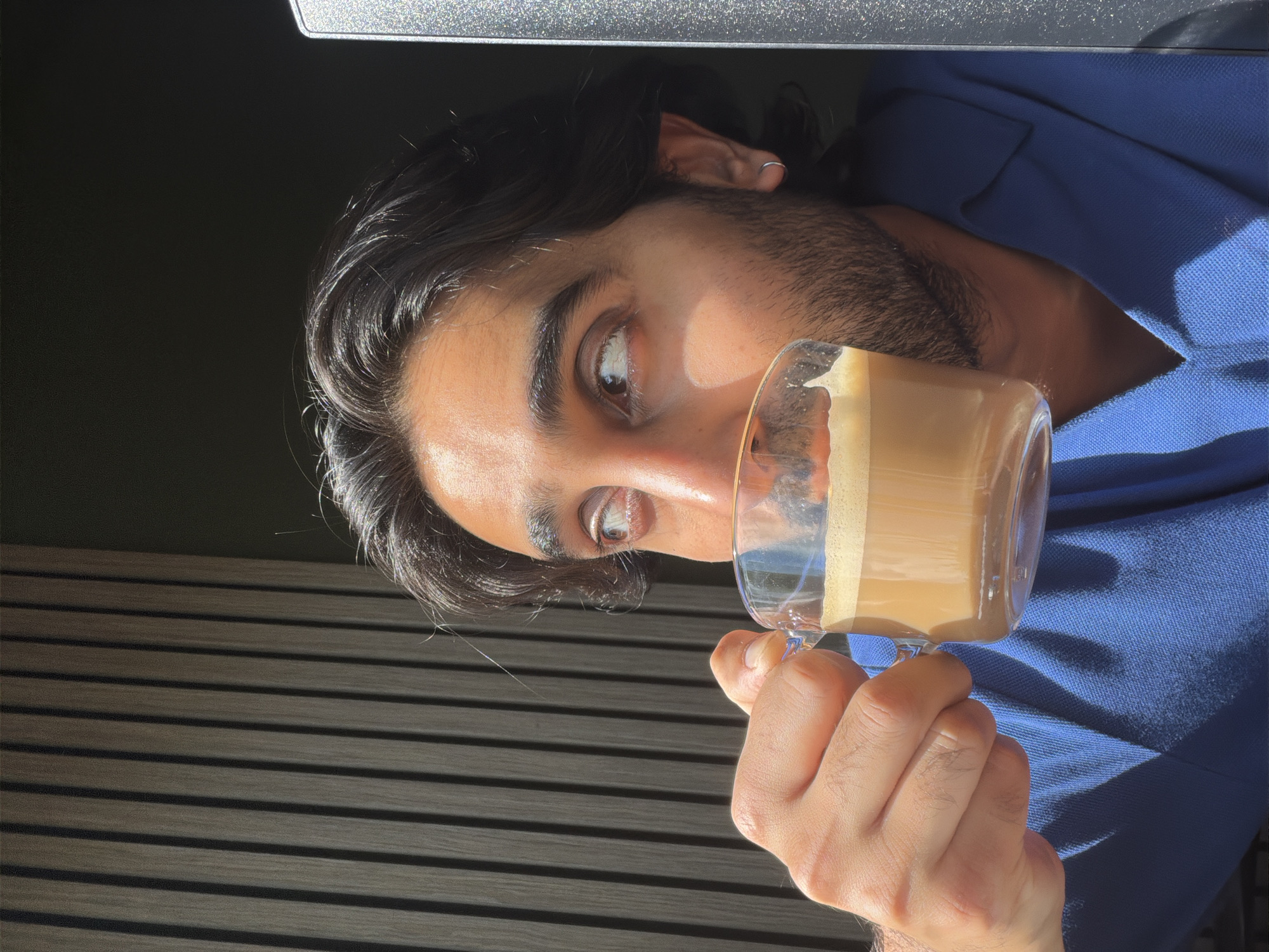The image shows a close-up of a young man holding a clear glass mug of coffee with frothed milk. He is wearing a blue collared shirt. The man has dark, wavy hair, dark eyebrows, and is looking towards the left side of the frame with a thoughtful expression. His right hand, with visible veins and slight stubble, is holding the mug. The background consists of dark wooden blinds and a dark surface above, suggesting an indoor setting, possibly by a window, with natural light illuminating the scene. A bright, speckled area at the top of the image hints at the presence of a screen or reflective surface. The lighting suggests it is daytime, with sunlight creating shadows. No visible text is present in the image.