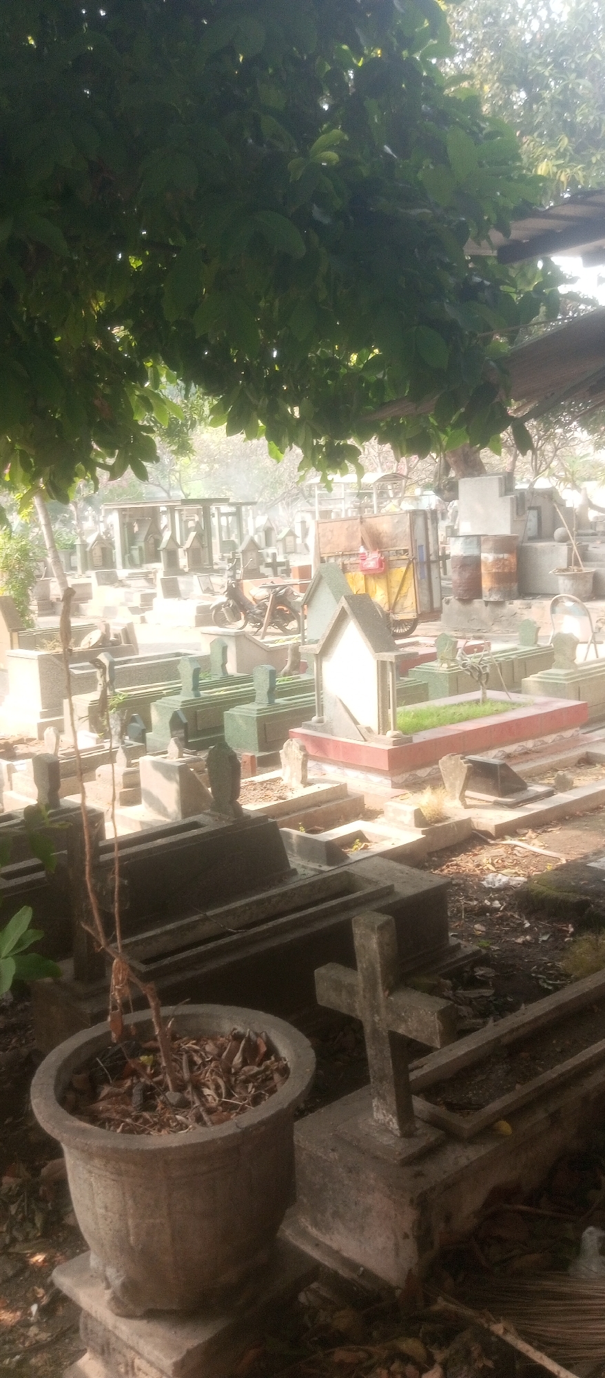 The image depicts a cemetery scene within a city, possibly Kediri City, Indonesia, characterized by numerous graves and tombs. In the foreground, a large, weathered concrete planter contains dry leaves and a thin, dead plant. To its right, a concrete tomb with a sloped top and a smaller, cross-shaped grave marker are visible. The background reveals a dense arrangement of various grave structures, some with intricate designs and small, house-like roofs. A motorcycle is parked among the graves, suggesting recent activity or a means of transport. Further in the background, a yellow cart with a wooden structure and several metal barrels stand near what appears to be a covered area or shed. Overhanging tree branches with green leaves cast shadows across the scene, indicating it is daytime. The overall impression is one of a well-established, albeit somewhat unkempt, burial ground.