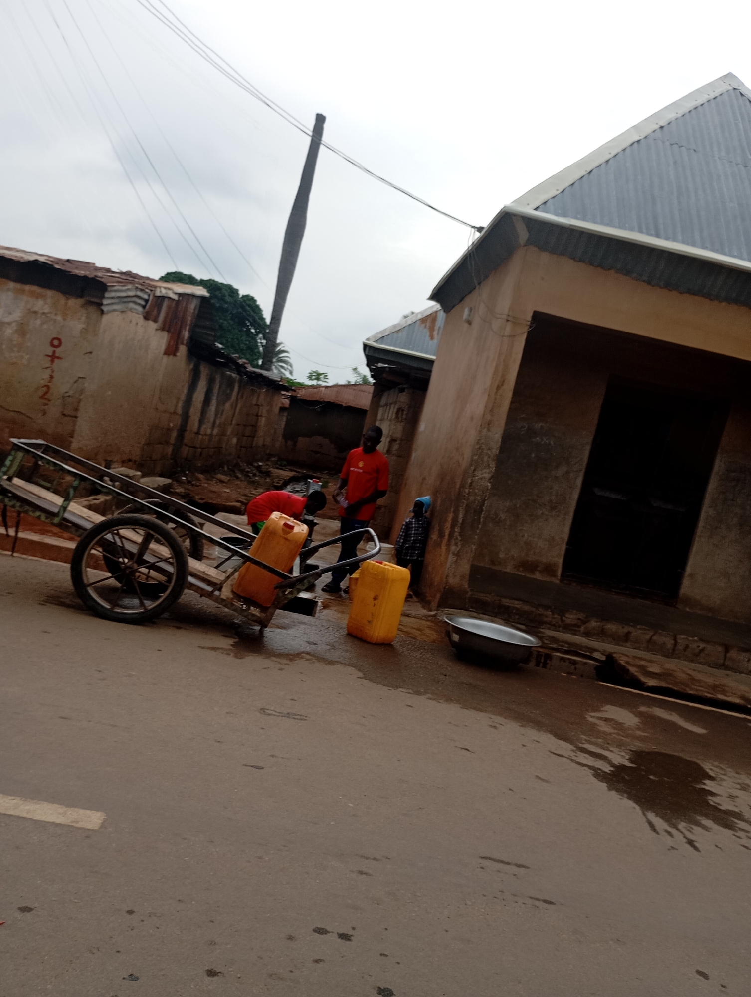 The image depicts a street scene in Shendam, Nigeria. In the foreground, a two-wheeled cart with a metal frame and wooden planks is parked on a paved road. The cart has one large wheel visible on the left side. Next to the cart, on the right, are two large yellow plastic containers, one upright and the other tipped on its side, with some water on the ground.
Several people are present. Two individuals appear to be interacting with the yellow containers. One person, wearing a red shirt, is stooping near the tipped container. Another person, also in a red shirt, stands to the right, facing away from the camera. A third person, wearing a plaid shirt and a blue head covering, is standing near the building on the right.
The environment consists of buildings with corrugated metal roofs and mud or stucco walls. Power lines are visible overhead, supported by a wooden pole. The ground appears wet, with puddles and water stains. The sky is overcast. The overall impression is of an outdoor urban or semi-urban setting, possibly involved in water collection or distribution.