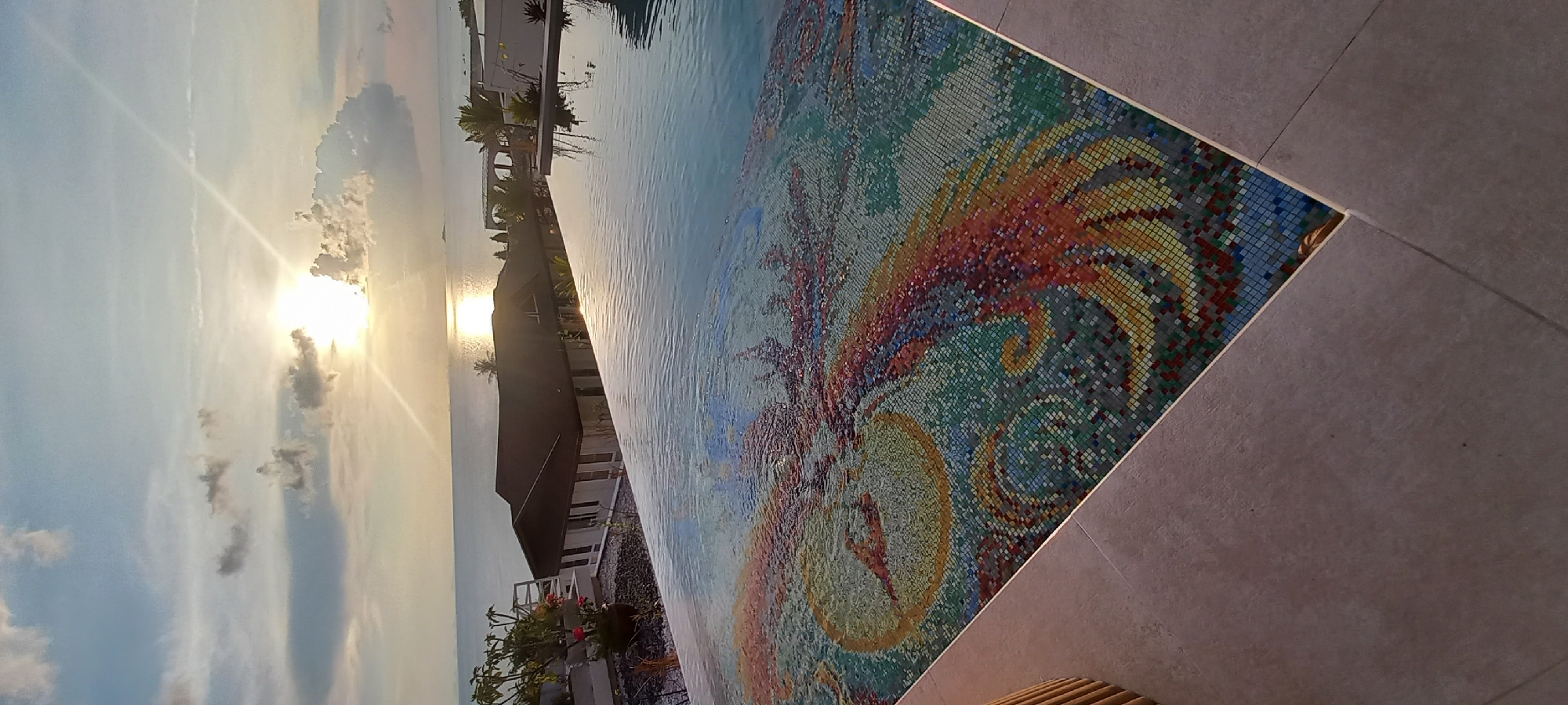 The image shows an infinity pool overlooking a sunset over water, likely the sea. The pool's edge is decorated with an intricate mosaic pattern featuring shades of red, orange, yellow, green, and blue. The sky is light blue with some clouds, and the sun is partially obscured by the clouds. A building with a dark roof is visible in the background, along with some palm trees and other vegetation. The setting appears to be Baan Wok Tum, Thailand, based on the presence of tropical vegetation and coastal scenery. The pool's water is reflecting the colors of the sky and the setting sun.