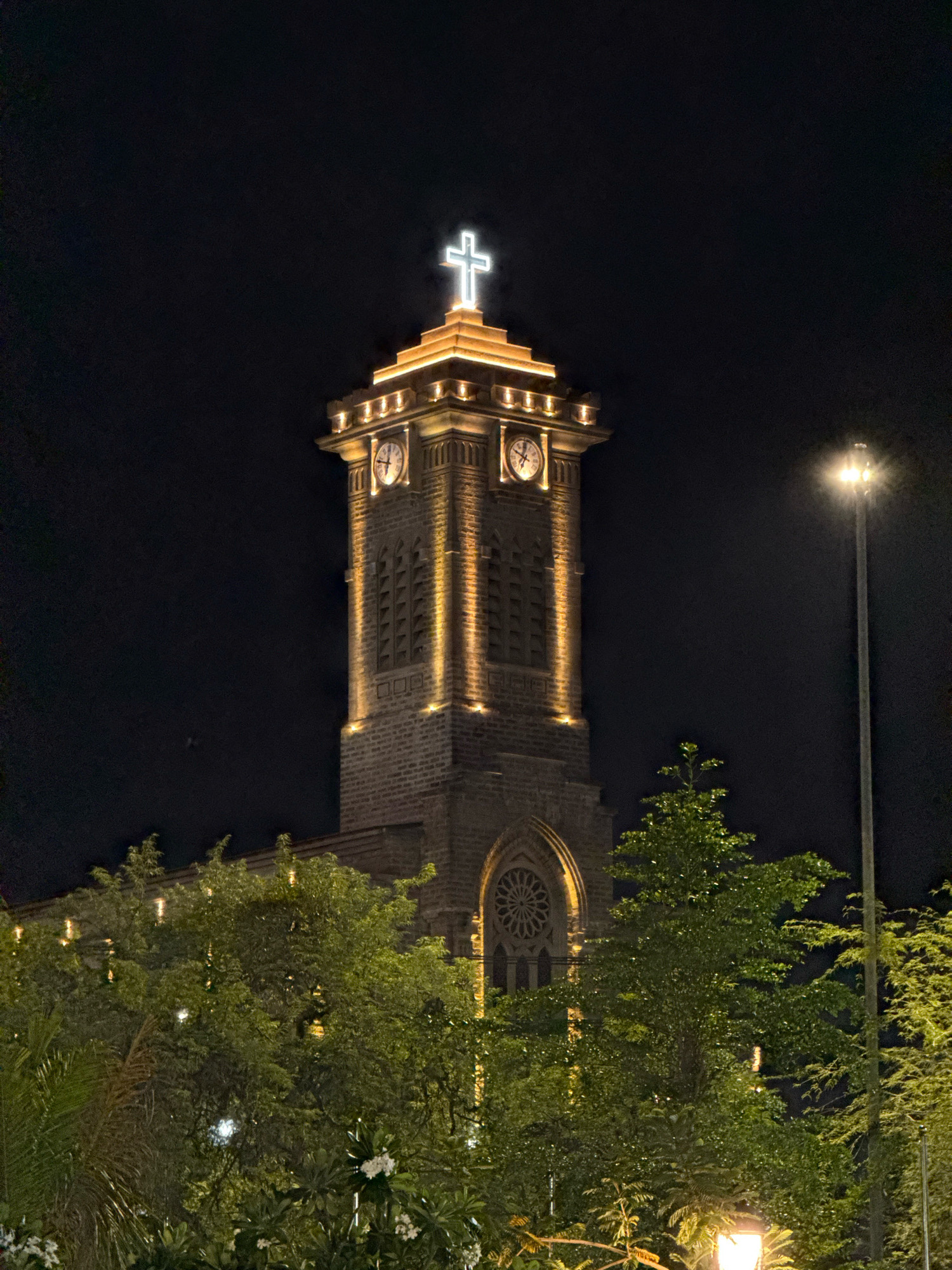 A nighttime exterior shot captures a stone church tower, illuminated by warm spotlights. The tower features two visible clock faces, ornate architectural details, and a bright white cross at its apex, which is also lit. In the foreground, lush green trees with scattered lights obscure the lower part of the building and the surrounding area. A tall streetlamp stands to the right of the frame. The scene appears to be in Nha Trang, Vietnam, likely St. Joseph's Cathedral, given the architectural style and prominent cross. There are no people visible in the image.