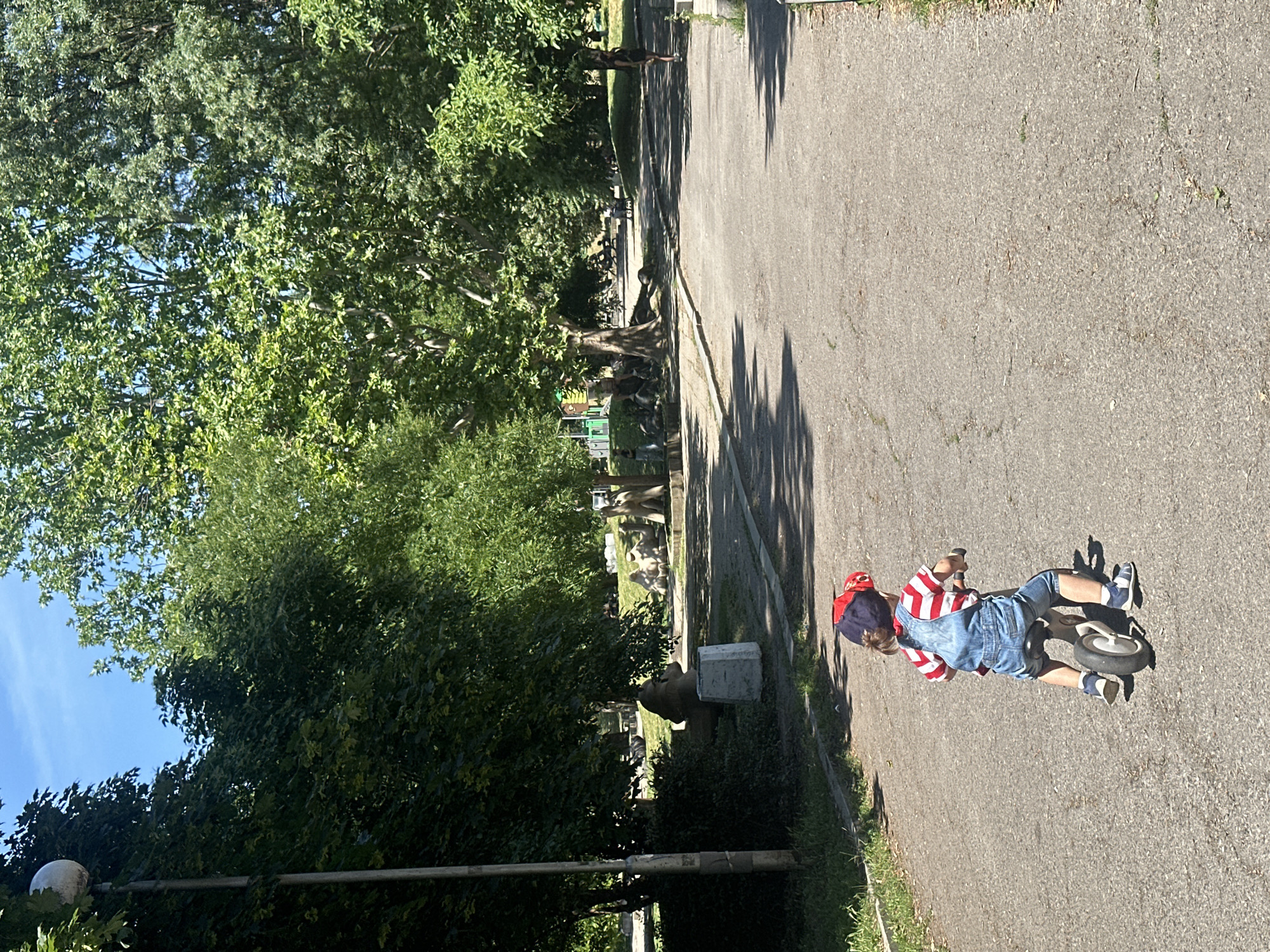 A child wearing a red and white striped shirt, denim overalls, a red and blue baseball cap, and blue shoes rides a balance bike down a paved path in a park. The path is lined with trees and grass, and in the background, there are playground structures and animal sculptures. The scene appears to be set in the Sofia Zoo, Bulgaria, given the presence of animal sculptures and the overall park-like environment. The sky is clear and blue, and the sun is shining, casting shadows on the path.