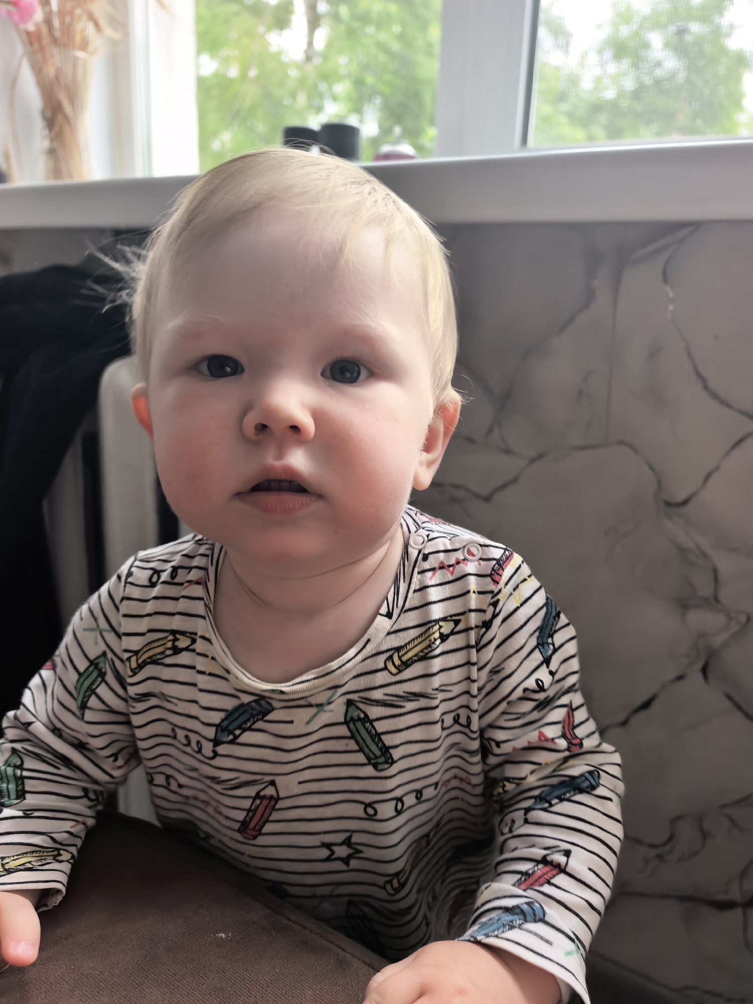 A close-up, eye-level portrait features a young baby with light blonde hair and blue eyes, looking directly at the camera with a neutral, slightly curious expression. The baby has fair skin and rosy cheeks. They are wearing a long-sleeved shirt with horizontal black stripes and a pattern of colorful crayons and small stars. The shirt also has visible popper fasteners on the shoulder.
In the immediate foreground, a brown, textured surface, possibly a high chair tray or table, is visible, with the baby's hands resting on it. The background is divided. To the left, behind the baby's head, part of a dark fabric object and a white window frame are visible. Further back, on the upper left, dried flowers or wheat stalks in a vase are partially seen.
A large window dominates the upper portion of the background, revealing lush green foliage and trees outside, suggesting an outdoor garden or park view. Bright natural light streams through the window, indicating it is daytime. To the right of the baby, a wall or partition features a distinct grey and white marbled or streaked pattern. On the window sill in the upper right, several dark, cylindrical objects are partially visible. The overall setting appears to be indoors, likely a home environment.