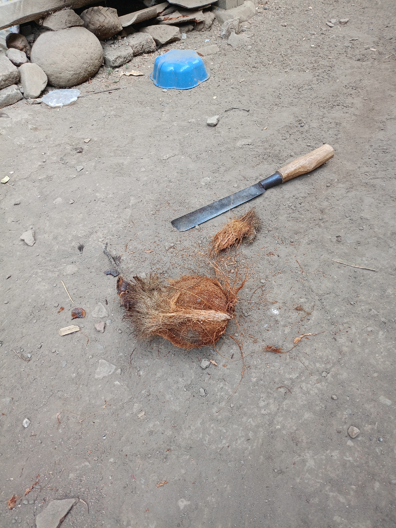 In Jelbuk, Indonesia, a scene unfolds on a gritty, earthen surface. A weathered coconut, its fibrous husk partially removed, lies center-frame, revealing a portion of its inner shell. Beside it rests a heavy-duty knife with a wooden handle, its blade showing signs of use. In the background, a collection of rocks and a discarded blue plastic basin are scattered across the ground. The environment appears to be an outdoor area, possibly a yard or an alleyway, characterized by its rough, unpaved terrain.