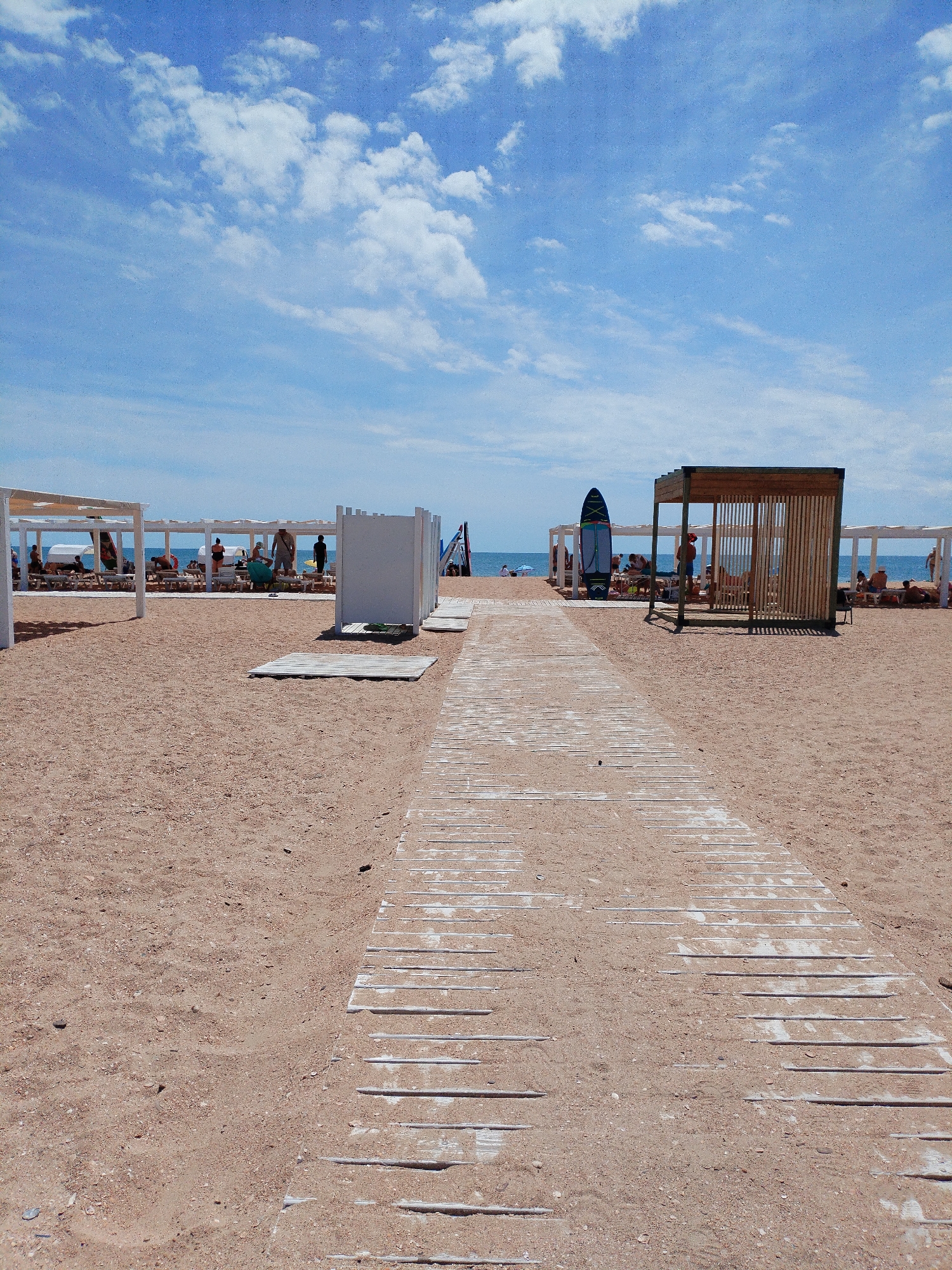 The image depicts a sandy beach with a wooden walkway leading towards the ocean. Several white structures, possibly sunshades or changing rooms, are present on the beach. A surfboard stands upright near one of the structures. In the background, people are visible relaxing on lounge chairs and walking near the water. The sky is blue with scattered clouds, and the sea appears calm. The scene suggests a typical day at a beach resort.