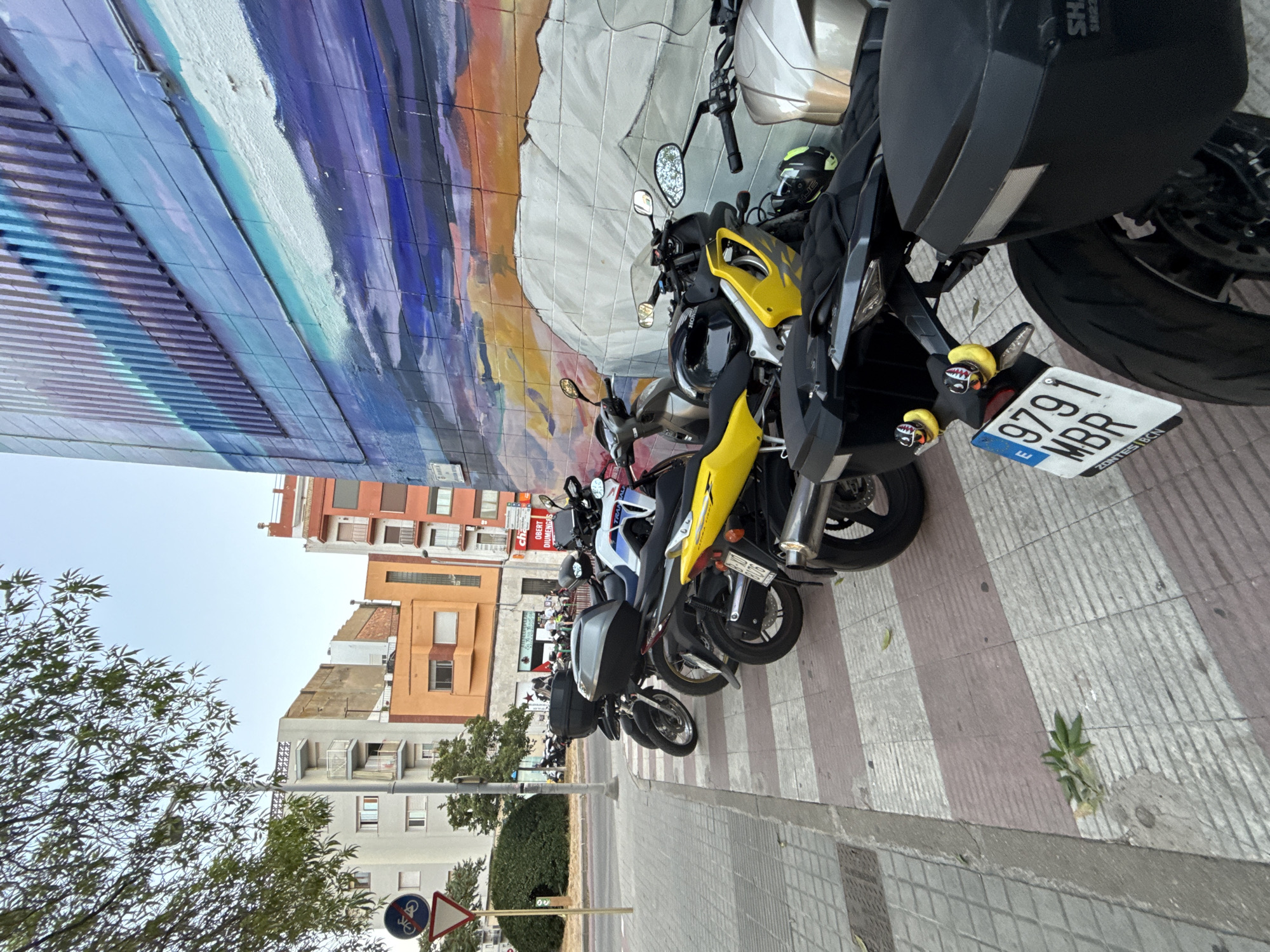 The image depicts a street scene in Blanes, Spain, with a row of parked motorcycles lining a sidewalk. The motorcycles are of various models and colors, including a prominent yellow sportbike and several scooters. They are parked on a checkered crosswalk. Behind the motorcycles, a colorful mural covers the side of a building, featuring abstract patterns and images of clouds and sky. In the background, several multi-story buildings with balconies are visible, along with trees and a street sign indicating a bicycle path. The sky is clear and blue. The perspective is from a low angle, focusing on the motorcycles and the mural. There are no people visible in the frame.