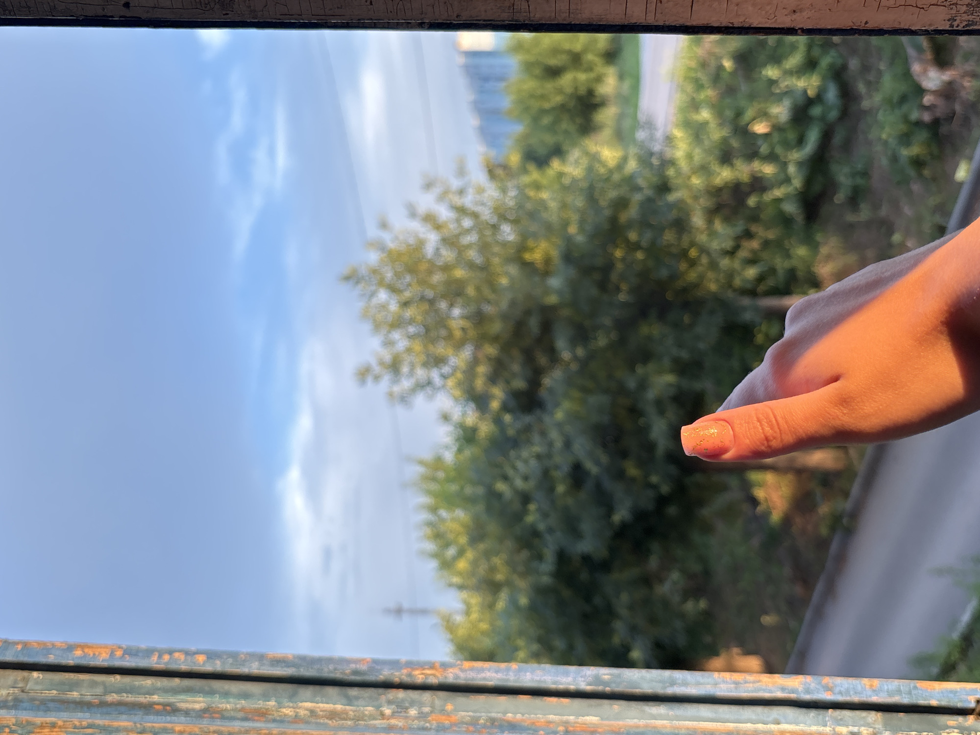 In the foreground, a right hand with a pinkish-orange hue from warm sunlight is extended upwards, with the thumb making a thumbs-up gesture. The thumb's nail is painted with a shimmery, light pink polish. The hand and thumb are positioned near an old, peeling window frame with visible rust and chipped blue paint.
In the background, a blurred landscape is visible through the window. Lush green trees dominate the mid-ground, with some lighter yellow-green foliage catching the sunlight. Beyond the trees, a road or path is faintly discernible, and in the far distance, several tall, blue-tinted residential buildings can be seen against a pale blue sky with wispy clouds. The scene suggests an outdoor setting, likely in a residential area of Inskoy, Russia, given the presence of apartment buildings. The overall lighting indicates it might be late afternoon or early morning due to the warm, directional sunlight.
