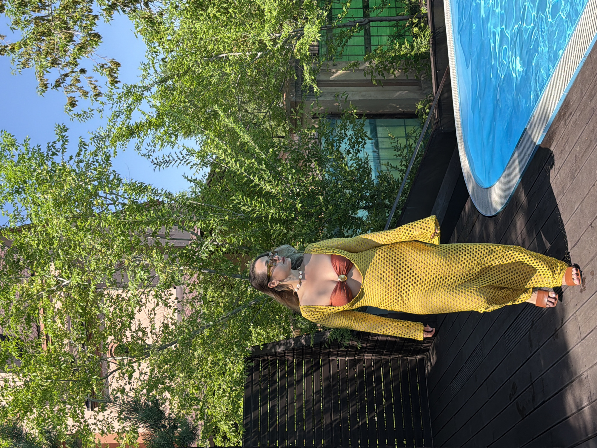 The image displays a woman standing on a dark brown wooden deck next to a bright blue swimming pool in Krasnoyarsk, Russia. She is wearing a long, sheer, yellow mesh-knit cover-up dress over a brown bikini top, which features a gold-colored ring detail at the center. She also wears brown flat sandals, sunglasses, and a pearl necklace.
The woman is positioned with her body slightly turned, looking towards her left. The setting is outdoors, bathed in bright sunlight, which casts some shadows on the deck. Behind her and surrounding the scene are abundant lush green trees, creating a natural and serene atmosphere. A building with green-tinted windows is partially visible through the foliage in the background, suggesting a residential or resort-like structure. The sky above is clear and blue. The scene conveys a relaxed, leisure activity on a sunny day. No visible text is present in the image.