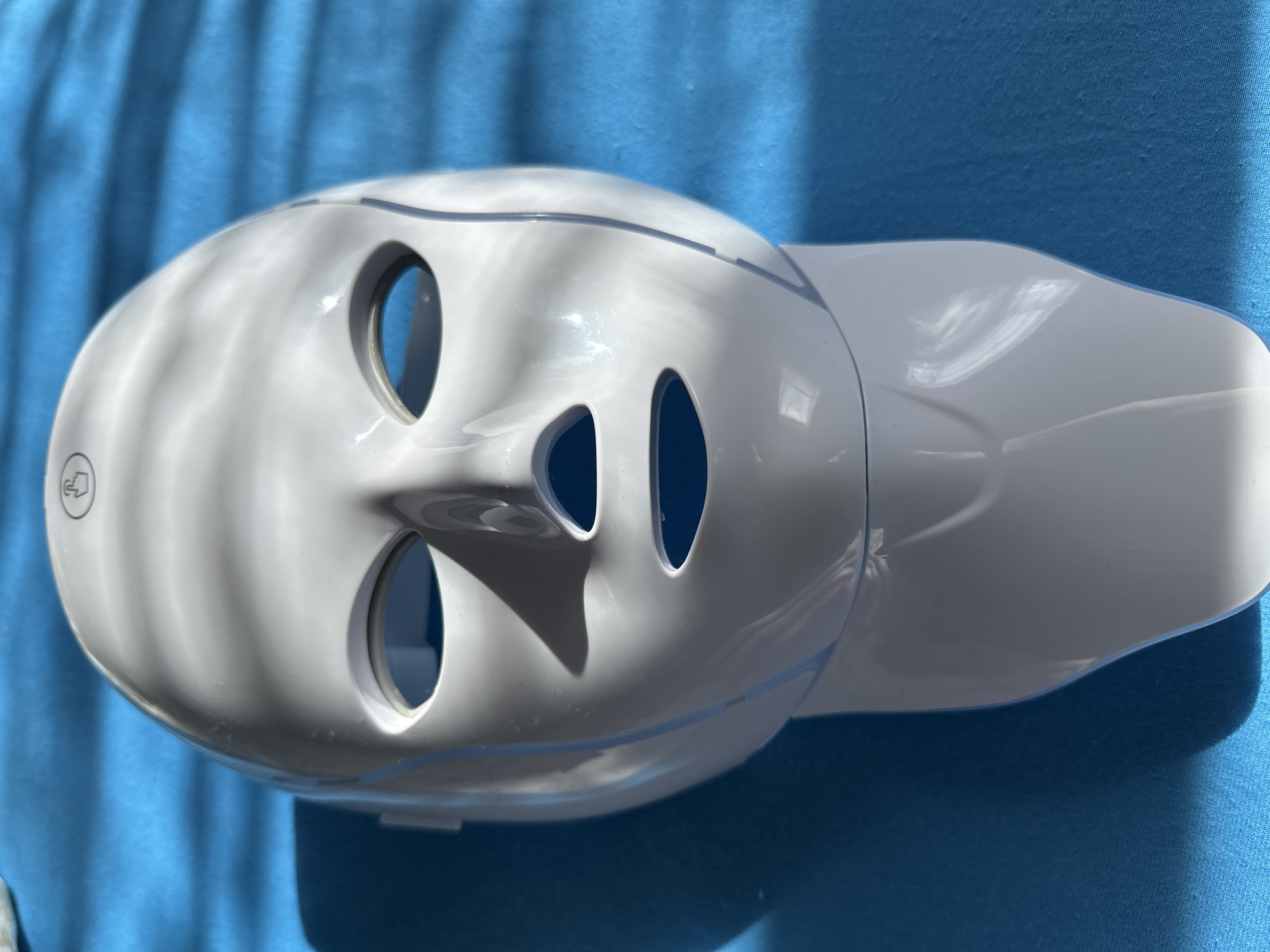 A white LED facial mask with eyeholes, a nose opening, and a mouth opening is displayed on a stand against a blue textured background. Shadows from venetian blinds fall across the mask, highlighting its contours. A small circular icon with a finger symbol is visible at the top of the mask. The mask is positioned on a white stand that resembles a neck. The image has been photographed from a slightly elevated, front-facing angle. There is no information in the image to suggest it is located in Podolsk, Russia.