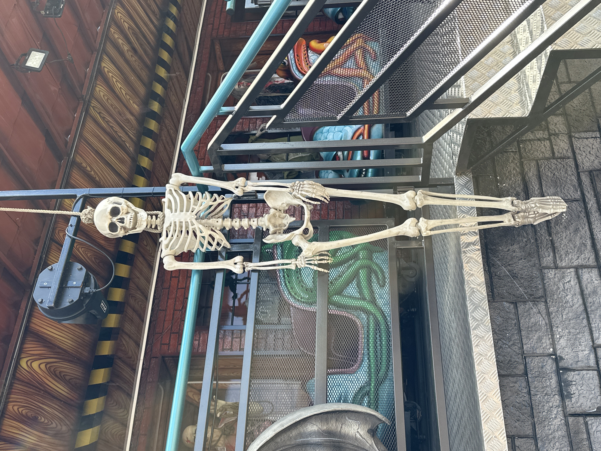 A hanging skeleton decoration, composed of plastic, is positioned in the foreground. The skeleton is suspended by a rope from an overhead structure that has a wooden-grain pattern and a black and yellow striped border. To the left and behind the skeleton, a carnival ride is visible, featuring a large grey wing-like structure and a green painted wall with a tentacle motif. Further to the right, metal stairs with black railings and blue supports lead up to the ride's entrance. On the ride itself, colorful figures resembling monsters or characters with large eyes and mouths are visible. The overall scene suggests a Halloween-themed attraction at a city fair or amusement park. The specific location in Tallinn, Estonia, is not identifiable from the provided image.