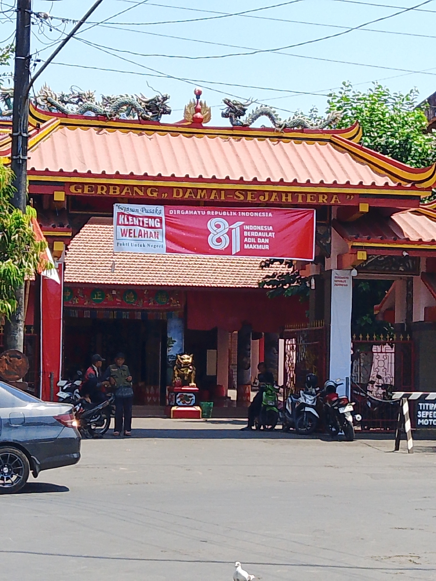 The image shows the entrance to Klenteng Welahan in Gedangan, Indonesia. The gate is adorned with dragon statues on the roof and features banners celebrating the 81st anniversary of the Republic of Indonesia. Two individuals are standing near motorcycles in front of the gate. A gray car is parked on the left side of the image. The environment appears to be a street, with trees and buildings visible in the background.