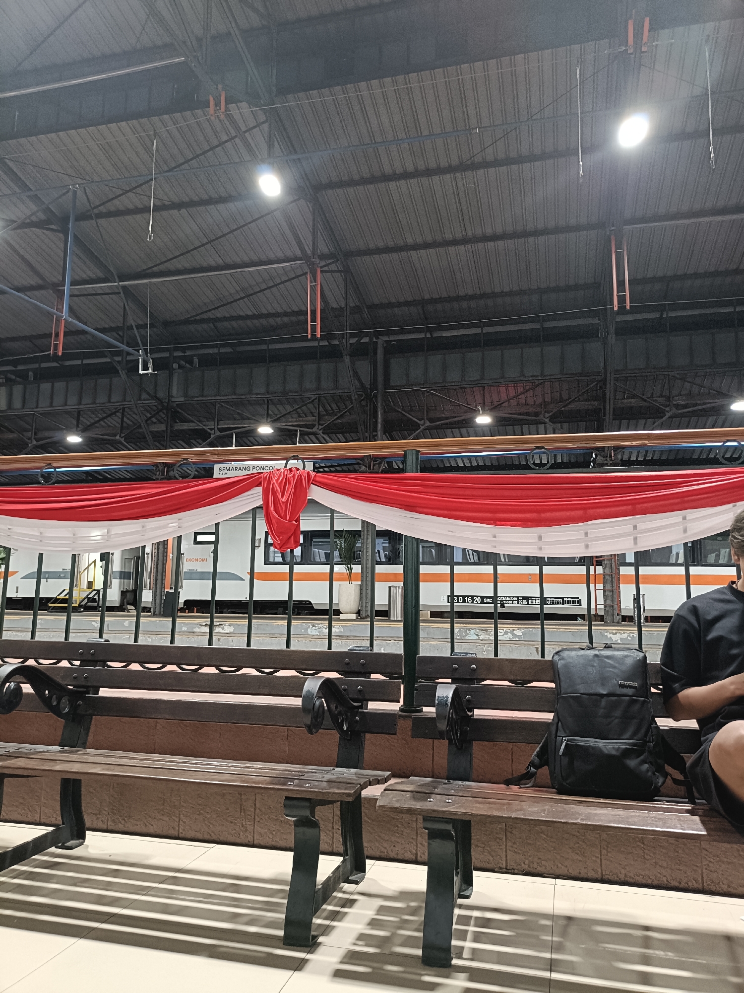 The image depicts a train station platform at night, likely in Semarang, Indonesia, indicated by the signage "SEMARANG PONCOL". The platform is covered by a large, industrial-style roof structure with visible beams and lighting fixtures. A train, predominantly white with orange and grey accents, is parked alongside the platform. Red and white draped fabric is strung across the platform, possibly for decoration or a national celebration.
In the foreground, there are several wooden benches with ornate dark metal legs. One bench in the foreground shows shadows from the lighting above, casting a striped pattern on the tiled floor. A black backpack rests on another bench. To the far right, a person, partially visible from the back, is seated on a bench, wearing a black t-shirt and shorts, and appears to be looking at their phone or a similar device. The overall atmosphere is one of a typical evening at a busy transportation hub.