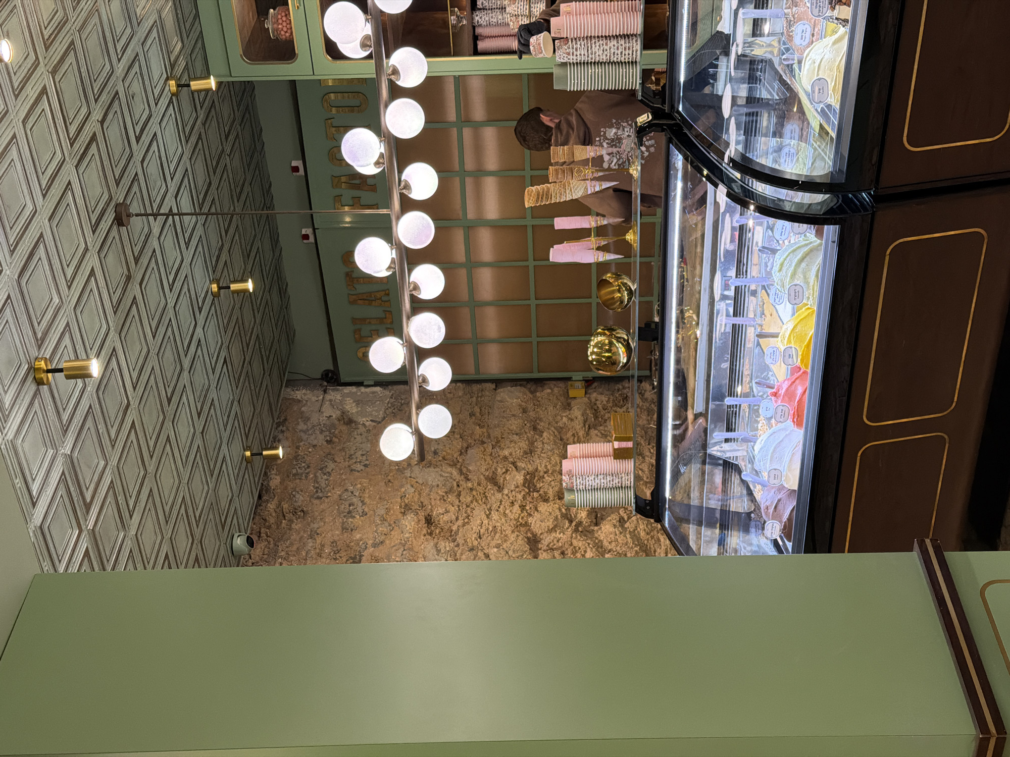 The image captures the interior of an ice cream shop in Tel-Aviv, Israel. A central feature is the illuminated display case filled with various flavors of gelato. A row of spherical lights hangs overhead. Behind the display, there's an individual preparing orders. Stacks of ice cream cones and cups are visible. The walls exhibit contrasting textures, one side with a patterned geometric design, and the other with a more rustic, natural stone finish. A security camera is mounted on the wall. The color scheme predominantly uses muted greens and browns.