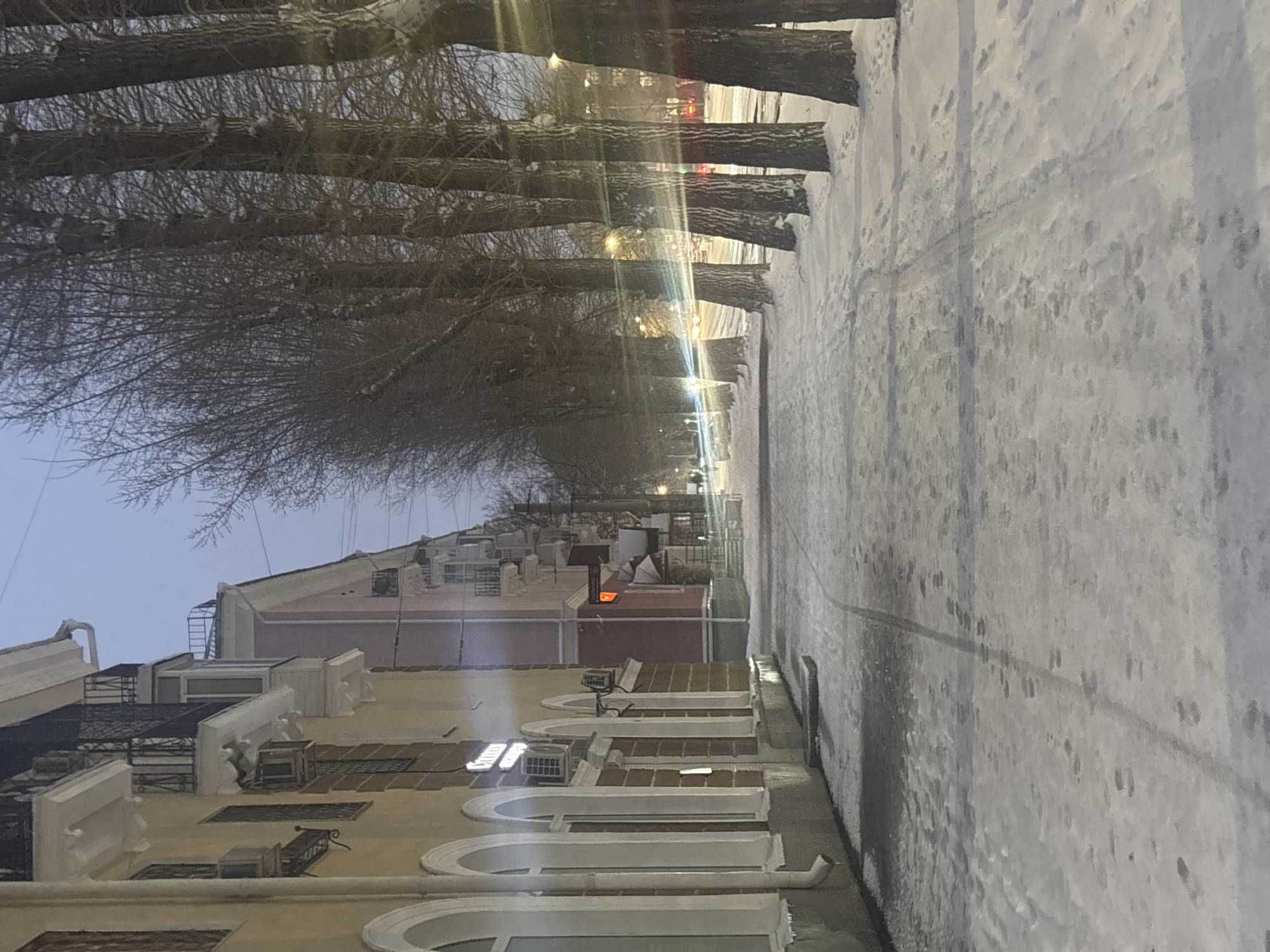 The image depicts a street in Perm, Russia, in winter. The street runs lengthwise, with buildings on the left and a snow-covered sidewalk on the right. Tall, bare trees line the street, their branches intertwining overhead. The buildings are multi-story, with architectural details such as arched windows, balconies, and what appear to be air conditioning units. A few lights illuminate the street, suggesting it is dusk or night. There's snow covering all surfaces, indicative of a winter setting. A person is seen walking far in the distance.