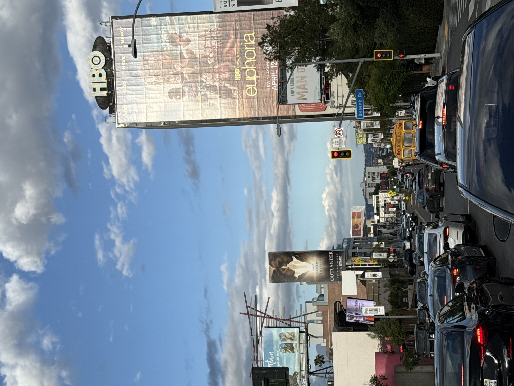 The image shows a street in West Hollywood, United States of America, densely packed with stationary traffic. Several large billboards are visible on either side of the street, advertising programs like "Euphoria" (HBO), "Man O Netflix", and "Outlander." A yellow school bus is among the vehicles stuck in traffic. The sky is partly cloudy with a mix of blue and grey. The street sign "Doheny Dr" is also visible.