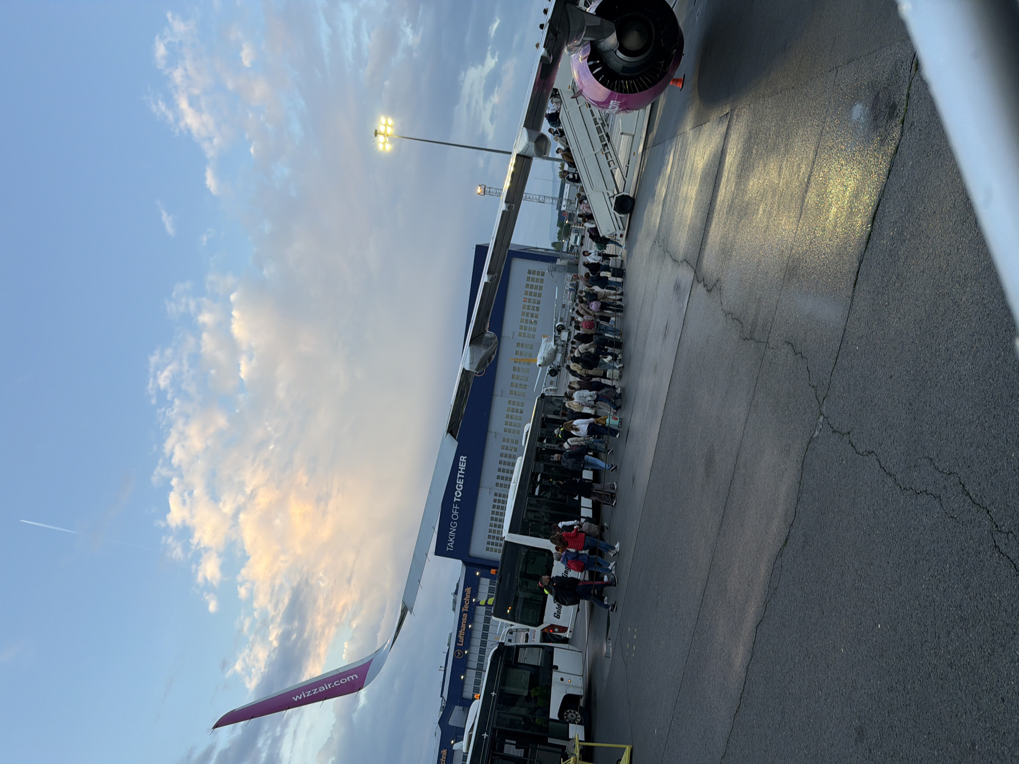 This image captures an active airport apron scene, likely during early morning or late evening due to the soft, diffused light and illuminated floodlights. The primary activity depicted is passengers boarding a Wizz Air aircraft.
A long queue of people is visible on the tarmac, waiting to ascend a mobile staircase into a purple and white Wizz Air airplane. The distinctive purple branding, including the text "wizzair.com", is clearly visible on the aircraft's wing and engine cowling.
Nearby, at least two white airport buses are positioned, with some passengers still on the ground near them, possibly having just disembarked from the buses or waiting for further transport. One bus shows "Gold Travel" written on its side. In the background, large airport buildings or hangars are present. One prominent building features the text "Lufthansa Technik" and "TAKING OFF TOGETHER" on its dark blue facade.
The ground is asphalt, displaying some cracks. Overhead, bright floodlights cast a yellowish glow. The sky is partly cloudy, transitioning from a clear blue to soft pinks, oranges, and greys, suggesting either sunrise or sunset. A faint contrail from another aircraft is visible in the upper left of the sky. The scene takes place at an airport in Sofia, Bulgaria.