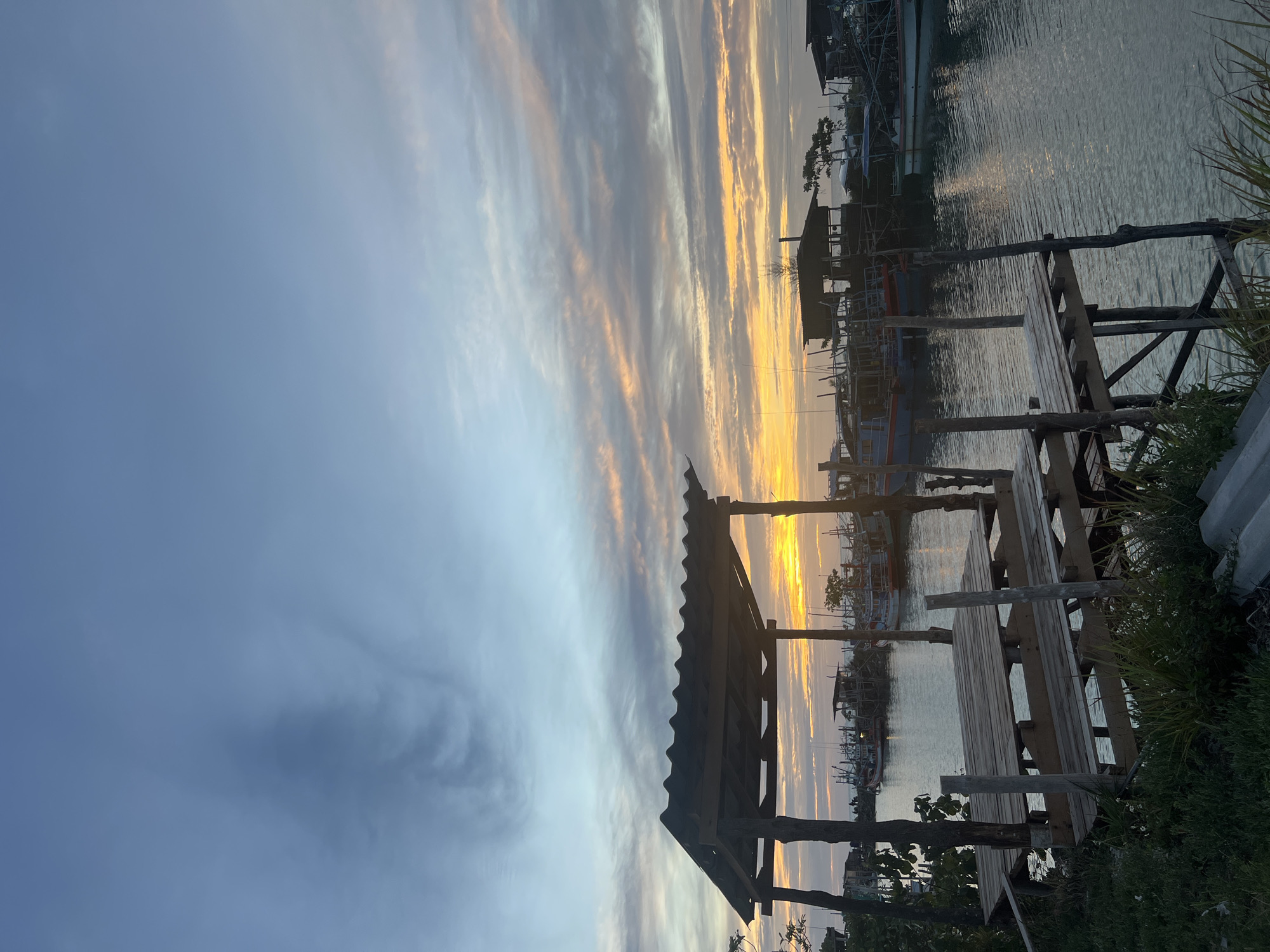 A vertical, medium shot captures a tranquil harbor scene at sunset. In the foreground, a rustic wooden picnic table with benches sits on a pier, partially shaded by a simple, corrugated metal roof supported by rough-hewn wooden posts. The pier extends over calm water, reflecting the soft twilight glow. Beyond the pier, a fleet of fishing boats is moored in the harbor, their silhouettes dark against the vibrant sky. The sky itself is a canvas of blues and grays, streaked with wisps of pink and orange as the sun dips below the horizon. The water is a deep, serene blue, with gentle ripples indicating a lack of wind. Lush green grass and foliage grow along the edge of the pier in the foreground. The overall atmosphere is one of peaceful serenity as day transitions into night.