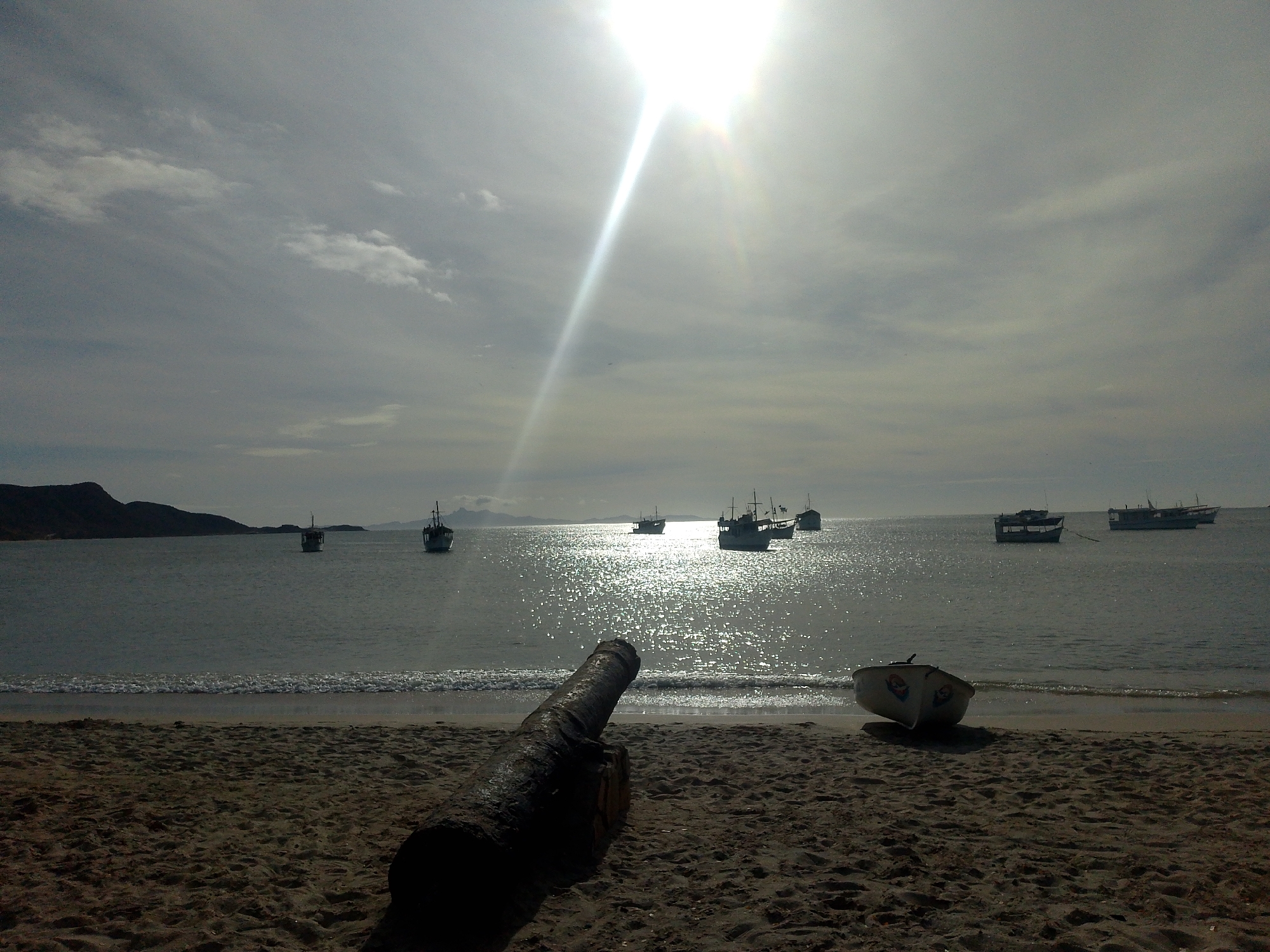 The image depicts a beach scene in Juan Griego, Venezuela. In the foreground is a rusty, old cannon pointed towards the water, along with a small white boat sitting on the sand. In the background, there are several fishing boats on the water. The sea reflects the sunlight, creating a bright, shimmering effect. The sky is overcast with the sun shining brightly, and a hint of land is visible in the distance to the left. The sand is textured, suggesting a relatively coarse grain. There are no people visible in the image.