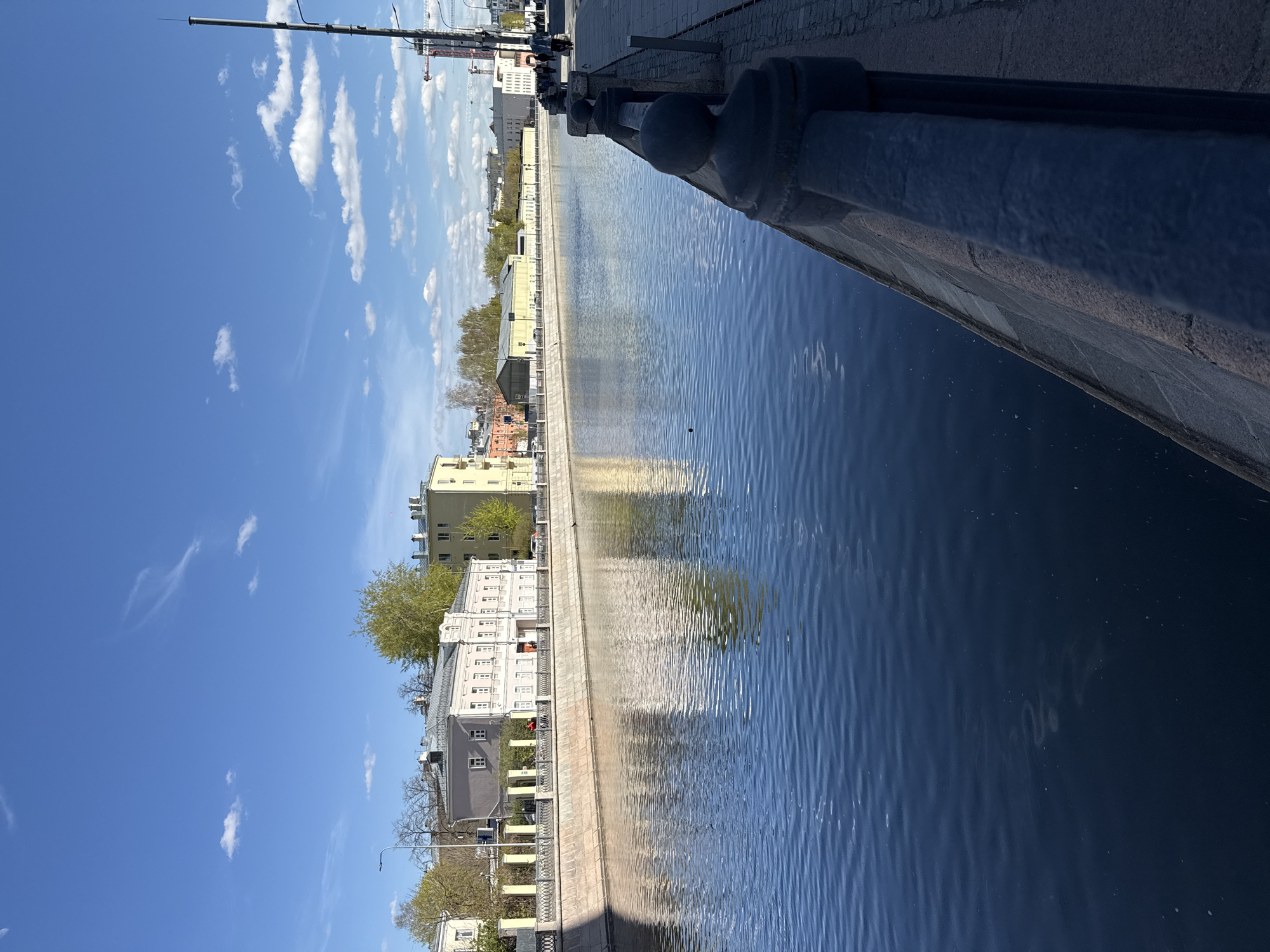 A photograph shows a canal in Moscow, Russia, lined with buildings and a stone embankment. The canal water is reflecting the buildings and the sky. The embankment is made of stone, and it has a metal railing along the edge of the canal. In the background, there are several buildings, including a high-rise under construction. The sky is blue with some white clouds.