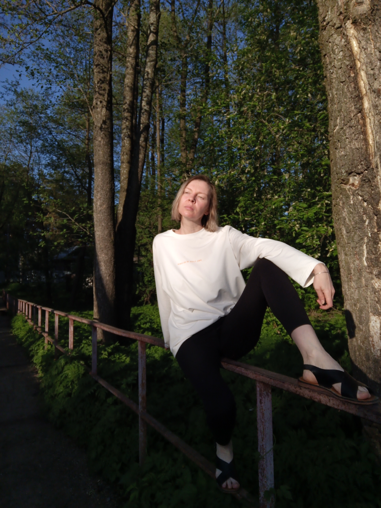 A fair-skinned woman with blonde hair is seated on a rusted metal railing in a park-like setting. She is wearing a long-sleeved white shirt and black leggings, with black sandals on her feet. Her left leg is dangling down, while her right leg is extended along the railing. She appears to be looking upwards into the sunlight, her eyes slightly closed.
Behind her are several tall trees, typical of a forest environment. The setting appears to be in a wooded area with a path running alongside the railing. The light suggests it is either early morning or late afternoon.
While the prompt asks for the location in Chaikovsky, Russia, there is nothing in the image itself that confirms this location. Therefore, it is not possible to verify the location using visual information alone.