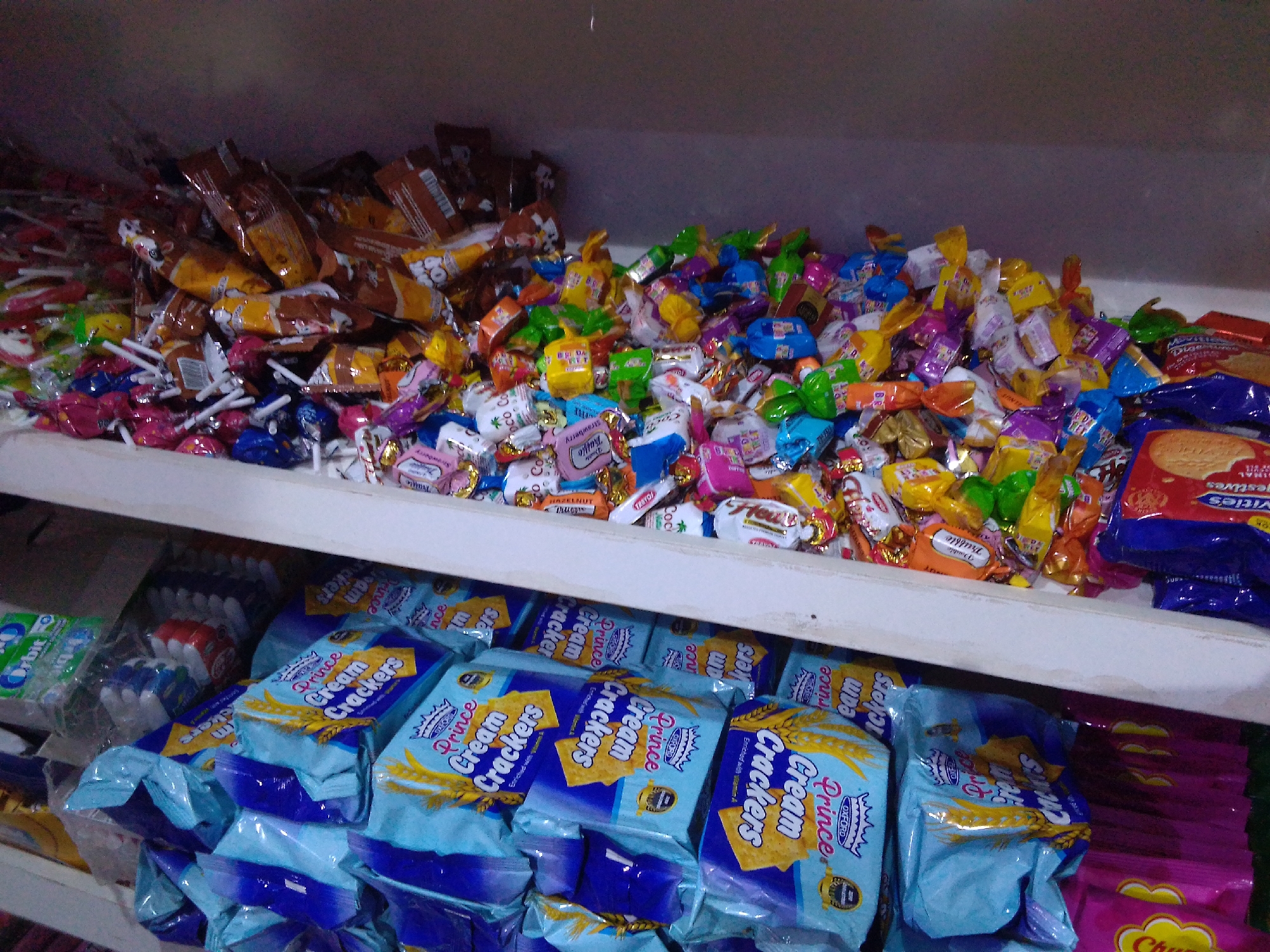 A shelf in a Nigerian store in Howayi, Nigeria, is filled with a large assortment of candies and packaged goods. The top shelf is overflowing with a chaotic jumble of individually wrapped candies in various colorful foils, including some chocolate-covered items, lollipops, and other sweet treats. The lower shelf is more organized, displaying several stacks of bright blue and yellow packages of "Prince Cream Crackers." To the left of the crackers, a green package of "Orbit" chewing gum and several smaller red and white packages are visible. Further to the right on the lower shelf, a large blue package with a picture of biscuits is prominent. The lighting in the store appears to be artificial, casting a somewhat dim glow on the products.