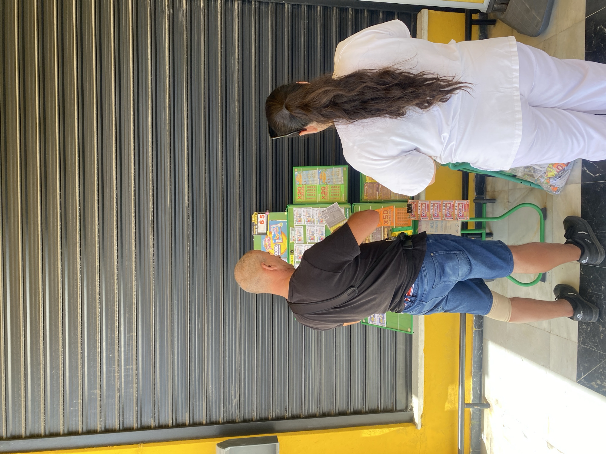 The image shows two people standing in front of a closed metal shutter. The person on the left is a man with a shaved head wearing a black t-shirt and blue denim shorts. He is looking down at something in his hands. He has a compression sleeve on his right leg. The person on the right is a woman wearing a white uniform. She has long, dark hair and is also looking down. They appear to be standing at a stand with lottery tickets and other merchandise. Some of the visible text on the merchandise includes "BOTE DE JACKPOT", "Verano", "0.000€", "ONCE A", "ILLO", "G24538", "ILUSIONATE", "DIO X50", "83520", "SANJUAN", and "x20". The scene suggests it might be daytime, given the lighting, and the setting appears to be an outdoor area, possibly a street or market stall.