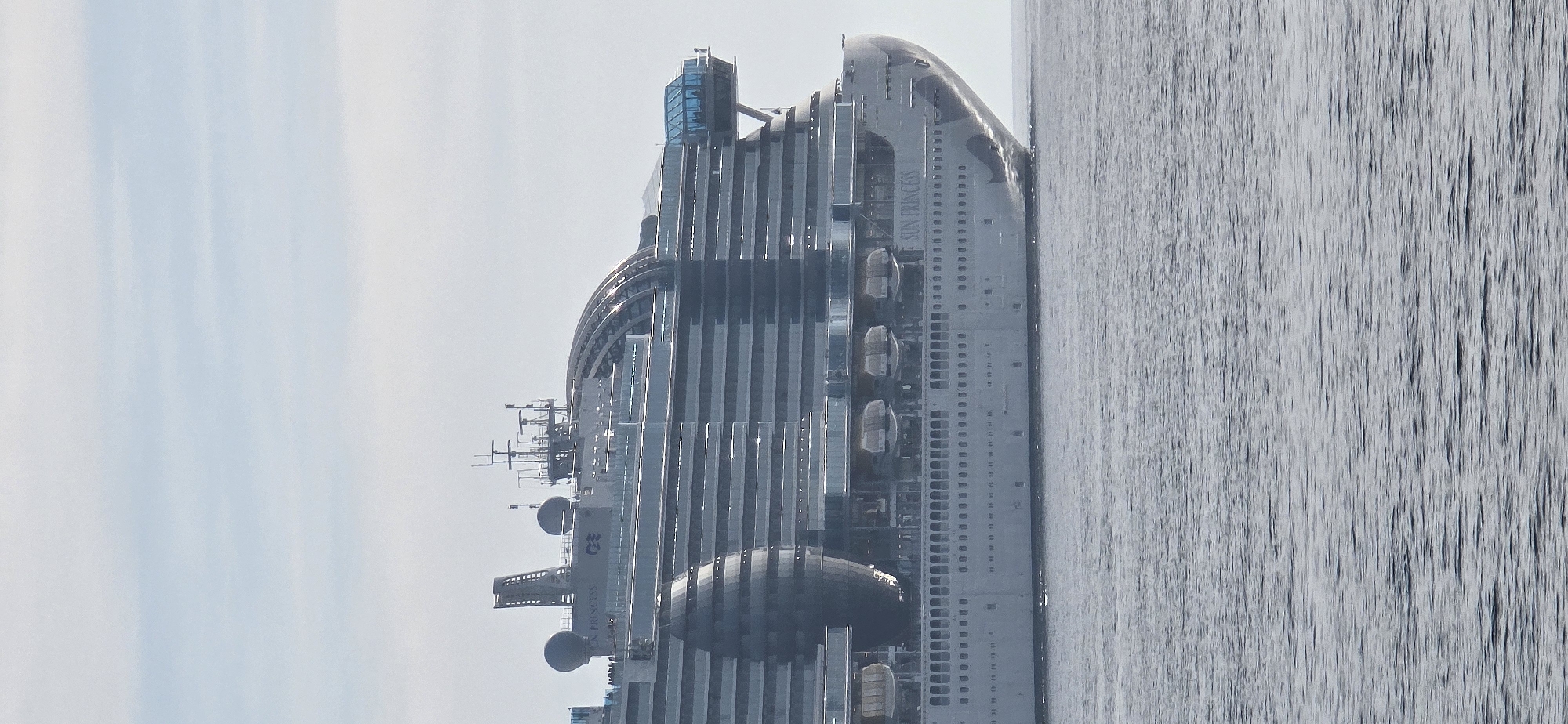 A large cruise ship, identified by the text "SUN PRINCESS" and the Princess Cruises logo on its bow, is depicted on calm water. The ship's bow is visible on the right side of the frame, rising out of the water. Its multi-deck superstructure features numerous balconies, several lifeboats mounted along the side, and prominent radar domes on its upper sections. The sky above is bright and appears overcast or hazy, providing soft, diffused light. The water surface shows slight ripples but is generally still. The scene is set during the daytime, and according to the provided context, the location is Bar, Montenegro.