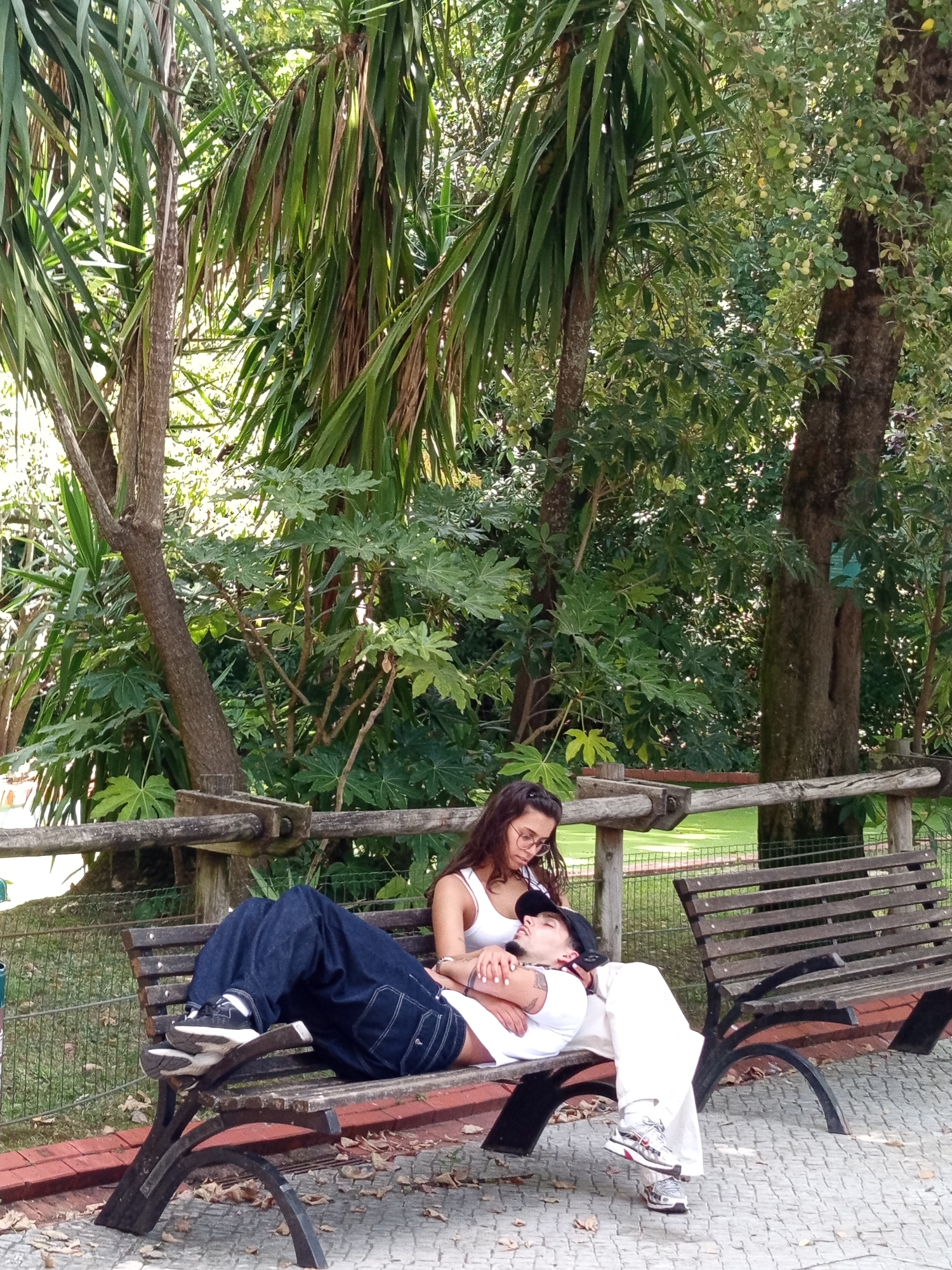 A young couple rests on a wooden bench in a park in Lisbon, Portugal. The man lies down, his head resting on the woman's lap, while she looks down at him. Both are dressed casually. The park is lush with greenery, including palm trees and other trees, with a wooden fence visible in the background. The ground is paved with stone tiles and scattered with fallen leaves. Sunlight filters through the trees, illuminating the scene.