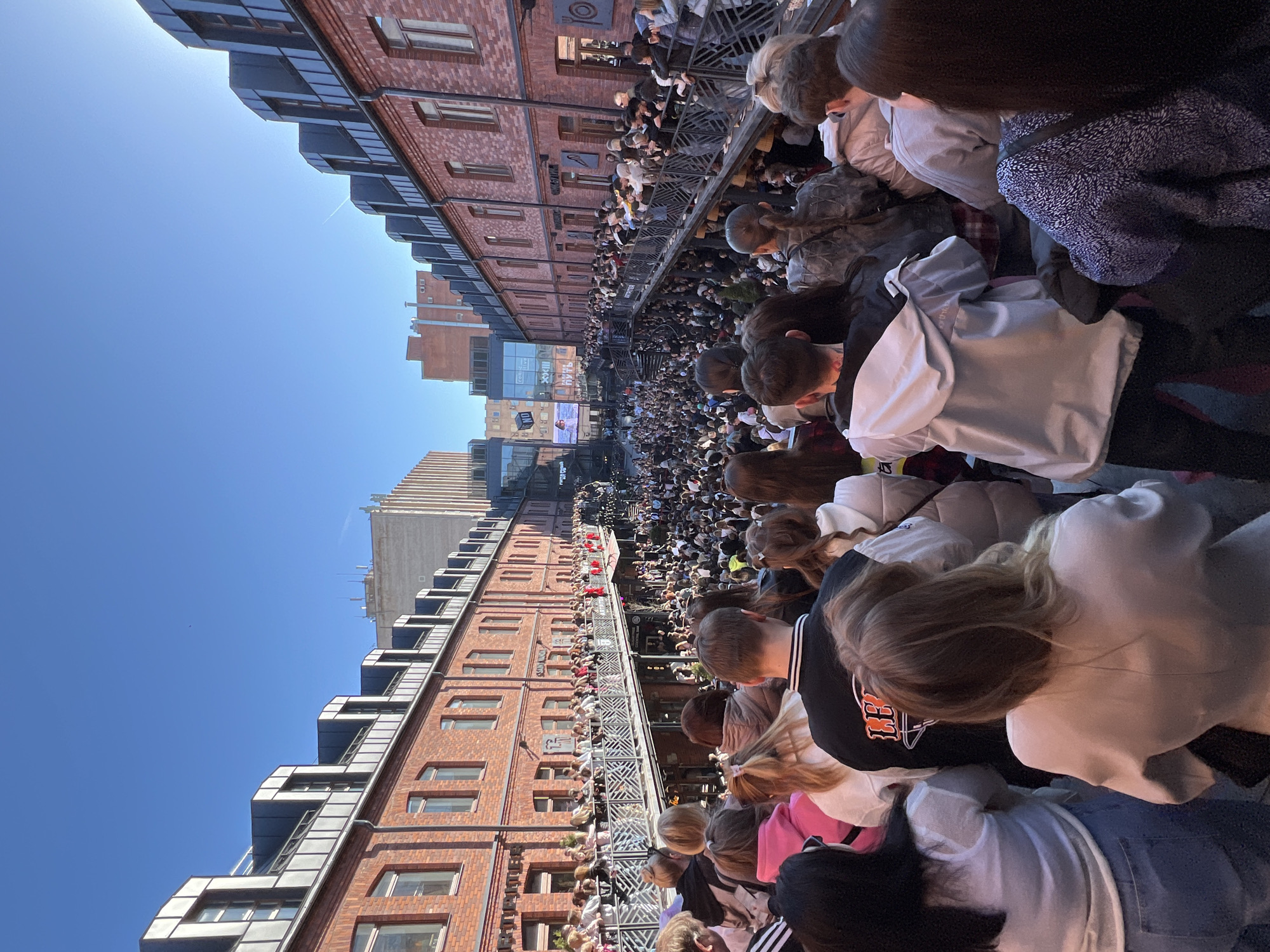 The image depicts a large crowd of people gathered in a narrow street between brick buildings. Many people are standing on balconies overlooking the street. The buildings have a modern industrial design. The scene appears to be taking place in St. Petersburg, Russia based on the Cyrillic writing on one of the buildings. A large screen is visible at the end of the street, suggesting a public event.