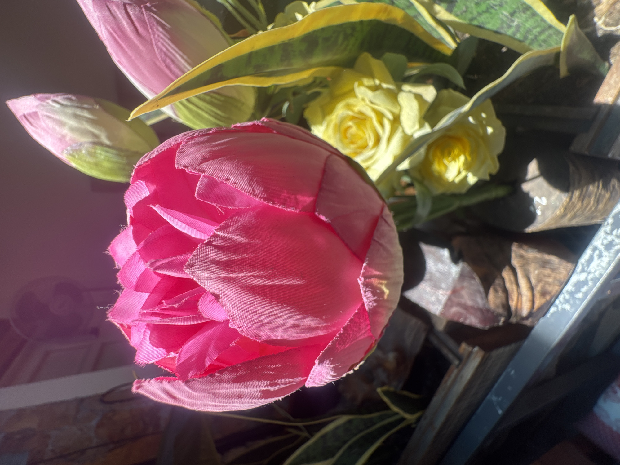 In a close-up shot of a floral arrangement, a vibrant pink, unopened tulip-like flower dominates the foreground, its textured fabric petals catching the light. Behind it, two delicate pink buds and a yellow rose peek through. The arrangement sits in a weathered wooden planter, partially obscured by a metal edge in the lower portion of the frame. A patterned wall and a ceiling fan are visible in the softly lit background, suggesting an indoor setting. This scene is located in the city of Bac Nha Trang Ward, Vietnam.