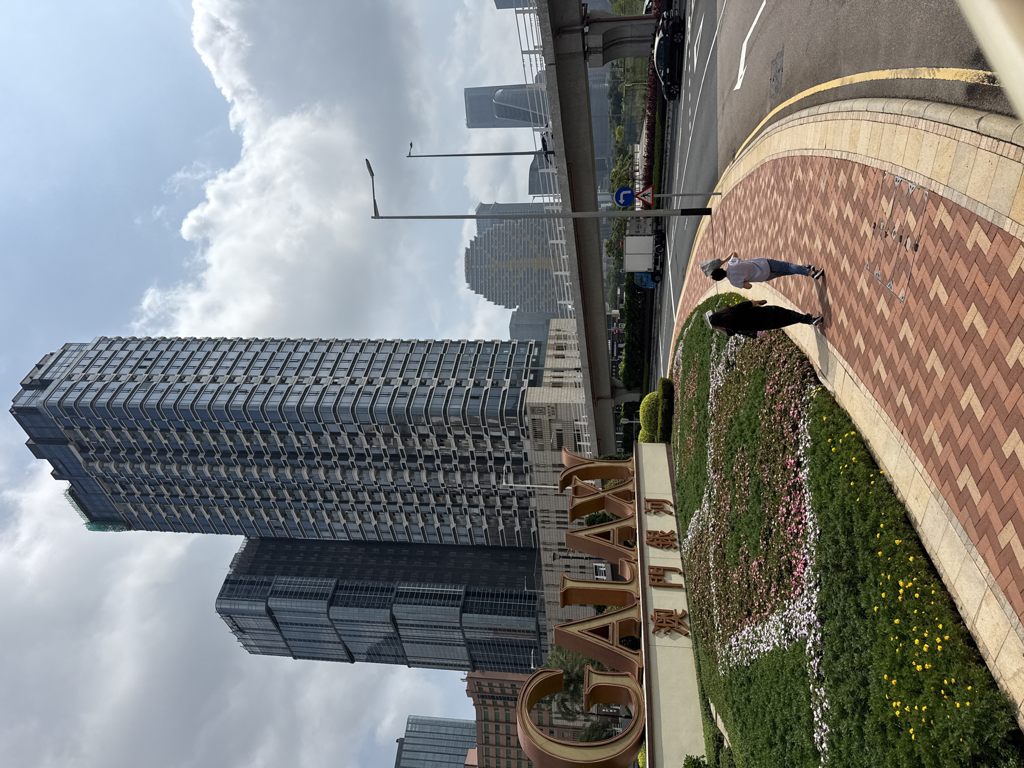 Captured from an elevated perspective, the image shows the Galaxy Macau complex in Macau, China. In the foreground, a flowerbed spelling out "GALAXY" borders a red-brick paved walkway where two individuals are walking. The architecture of the Galaxy complex, including a tall, rectangular, mirrored building, dominates the mid-ground. An elevated highway with vehicular traffic is visible to the side. The sky, filled with a mix of clouds and blue sky, serves as the backdrop. Street lights are also visible along the road.