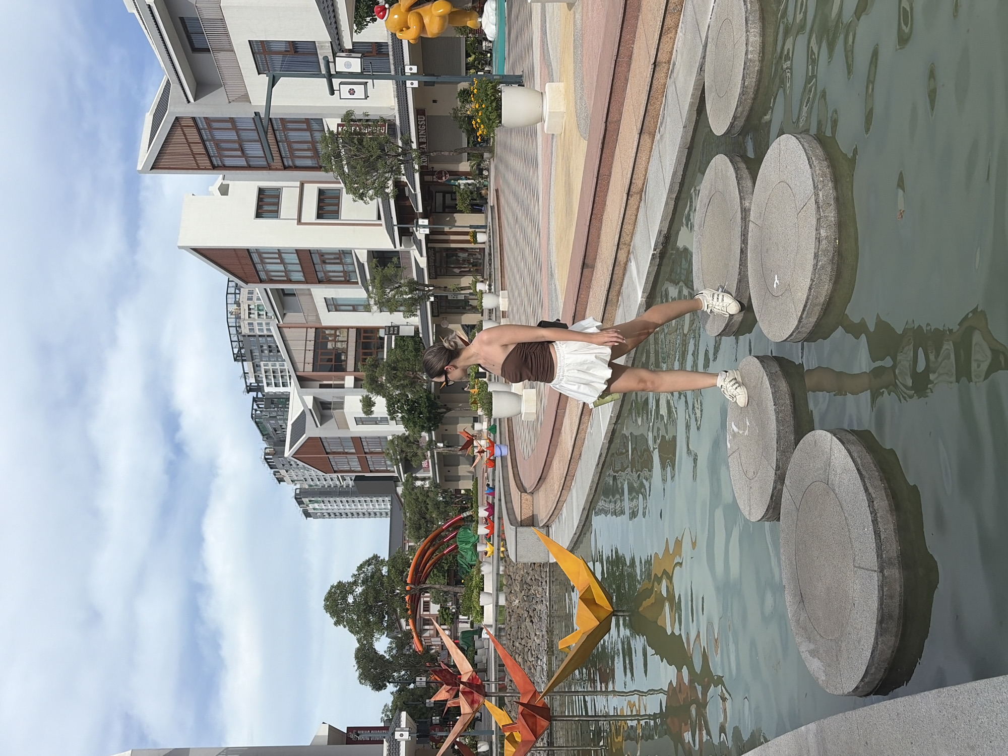 A woman is walking on stepping stones in a shallow pond in Nghĩa Trụ Commune, Vietnam. She is wearing a brown strapless top and a white skirt, with white sneakers. The stepping stones are round and gray, and they are placed in a pattern across the pond. In the background, there are modern buildings with a mix of white and wood-toned facades, and tall apartment buildings. There are also decorative paper cranes in the water, and a yellow sculpture of a bear wearing a Santa hat. The sky is cloudy.
