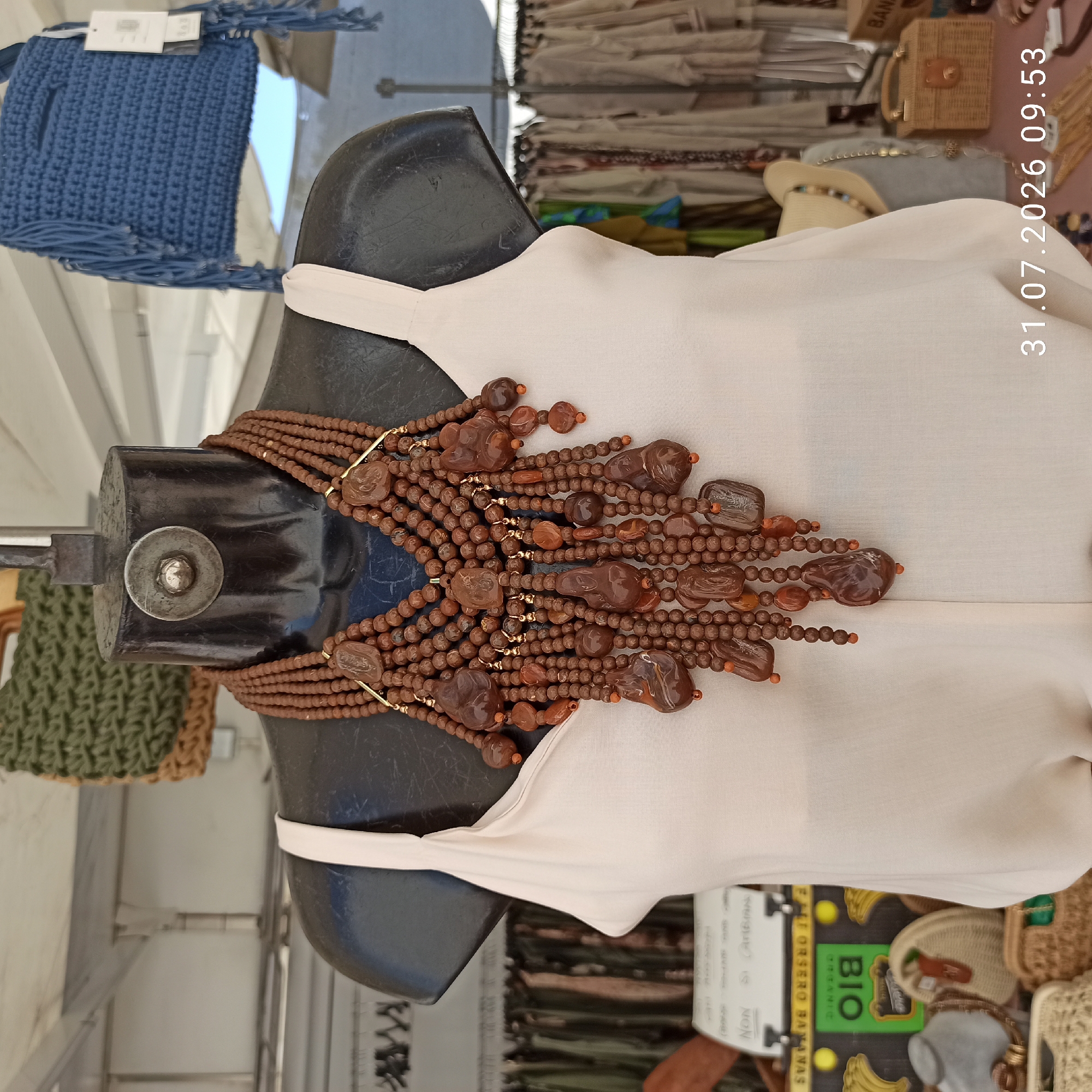 A dark-colored mannequin with a white tank top is displayed in front of a market stall. The mannequin is adorned with a large, multi-layered brown beaded necklace featuring carved brown pendants and gold accents. Behind the mannequin, various market items are visible, including racks of clothing, a blue crocheted bag with fringe hanging from the ceiling, and woven baskets. A sign with the word "BIO" and text indicating "ORGANIC" is partially visible on the left. The ground appears to be a pinkish-red surface. The scene is outdoors, likely at a street market or fair. A timestamp "31.07.2026 09:53" is overlaid on the bottom right of the image. The location is identified as Modugno, Italy.