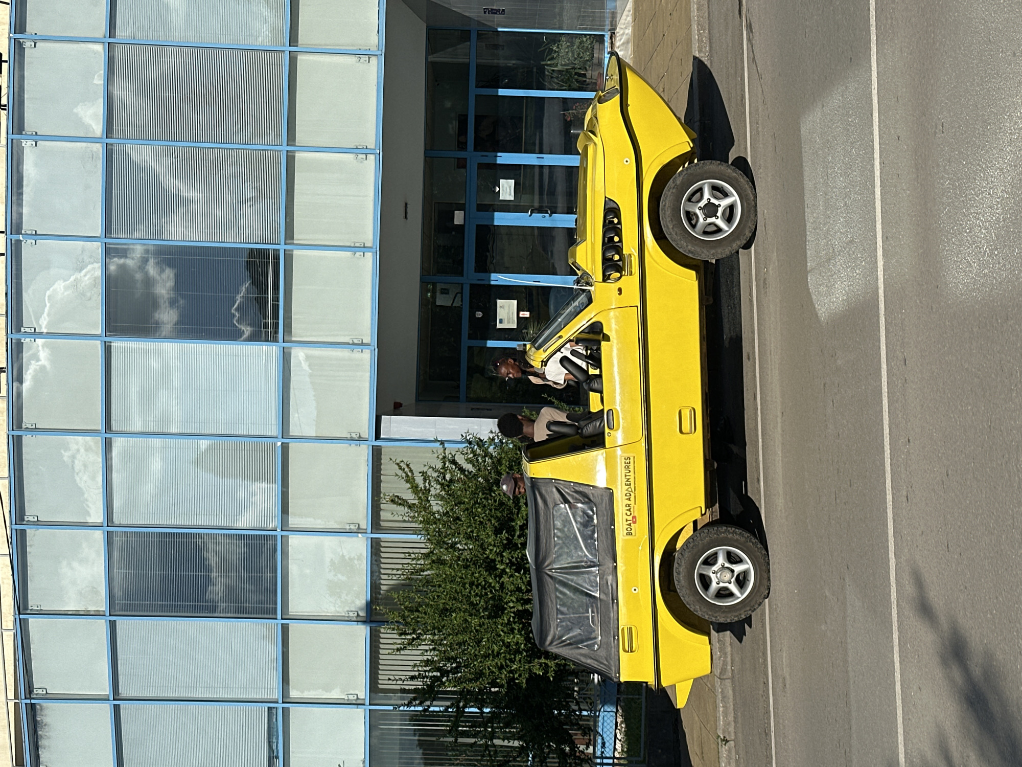 The image shows a yellow amphibious vehicle, identified by the text "BOAT CAR ADVENTURES" on its side, parked on a street in Gorna Oryahovitsa, Bulgaria. The vehicle has a black convertible top and appears to be a recreational vehicle. Three individuals are visible inside or near the vehicle. Behind the vehicle is a modern building with large, blue-framed windows that reflect the sky and clouds. The street in front of the vehicle has lane markings. A green shrub is to the left of the building. The scene suggests a sunny day.