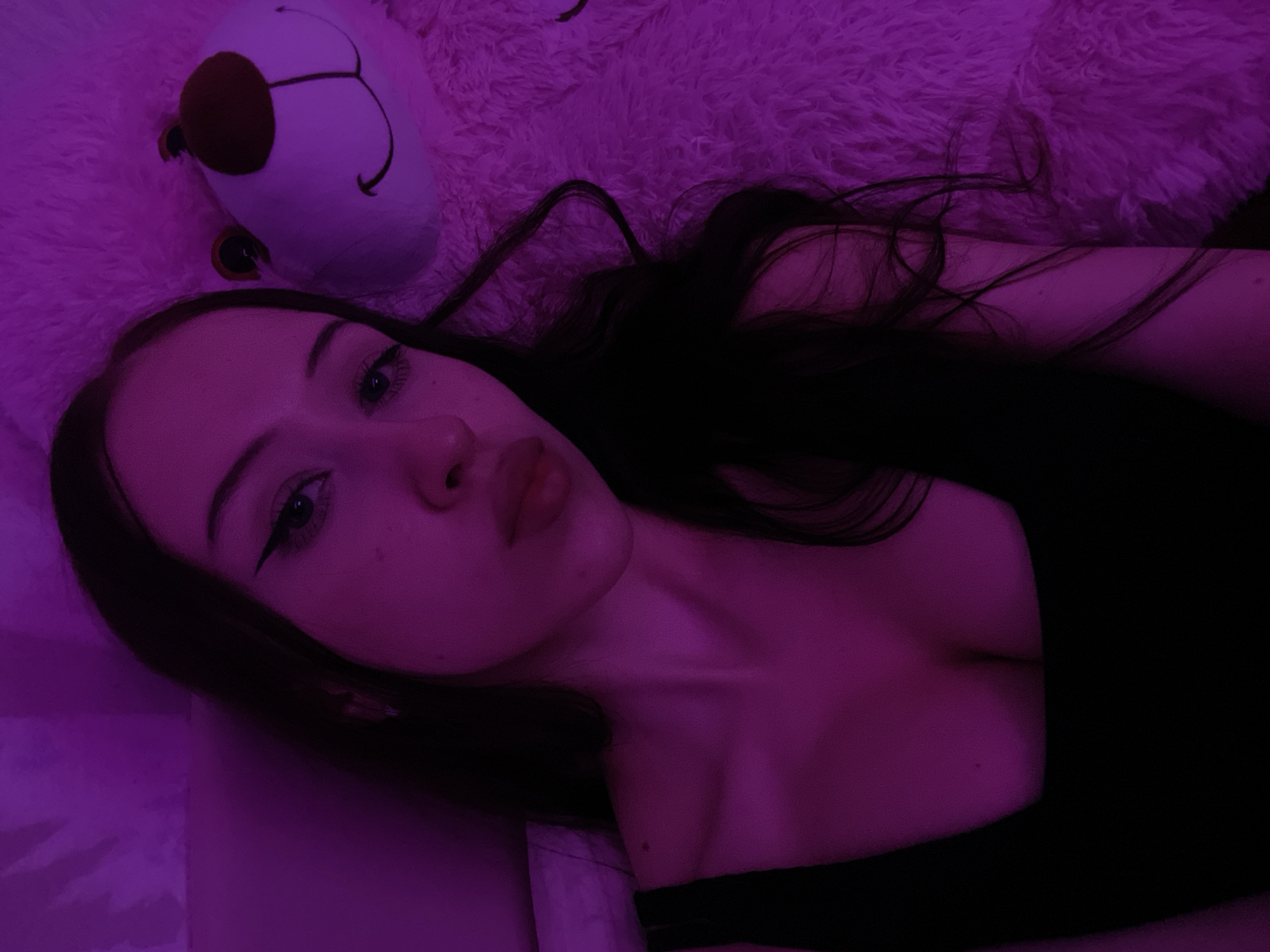 A young woman with long, dark hair is lying down, taking a selfie. She is wearing a black tank top. To her right is a large, fluffy, light-colored teddy bear with a brown nose patch. The scene is illuminated with purple lighting, casting a purplish hue over everything. The background appears to be a wall with a textured surface. There are no identifiable landmarks or contextual clues in the image that would indicate a specific location within the city of Votkinsk, Russia.