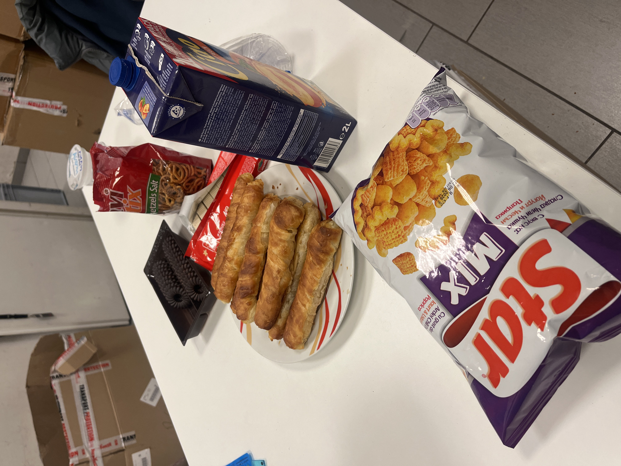 The image shows a tabletop with several food items. There's a package of "Star Mix" chips, a box of juice labeled "2L", a bag of "Mix Pretzels Salt", a plate with baked rolls, and a black container. Some of the text on the chip bag is in Bulgarian, and other text is in Romanian. There are boxes in the background. The text visible on one of the boxes includes the words "TRANSFORM FOR PROTECTION" and "SPORT". The origin of the food is Bulgaria. The location is Sofia, Bulgaria.