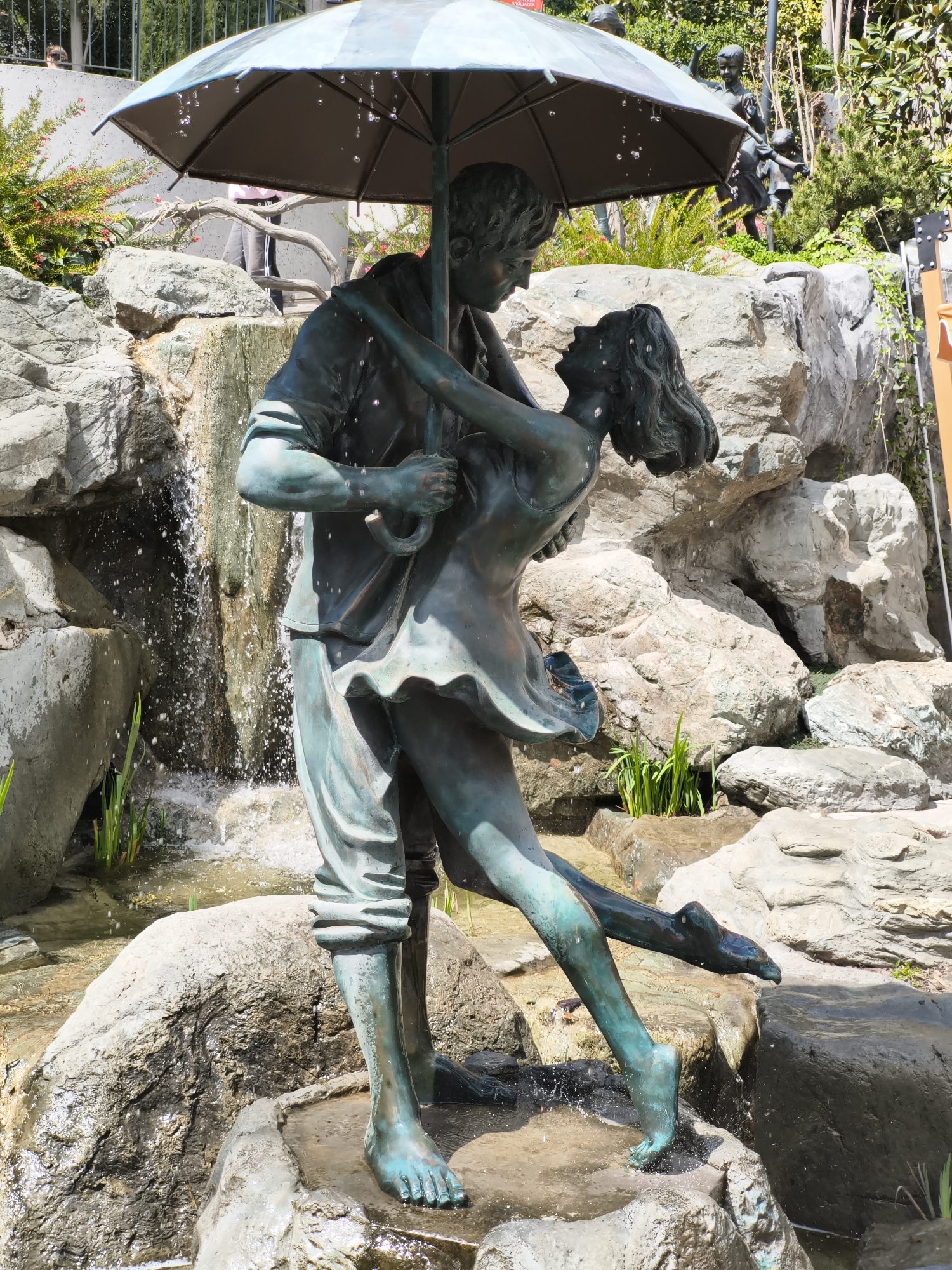 A full shot depicts a bronze sculpture of a couple dancing under an umbrella in an outdoor setting. The statue, which seems to be a fountain, is surrounded by various-sized rocks and vegetation. Water droplets are falling from the umbrella and the couple's bodies. Behind the statue, there is a rocky wall and more greenery, and in the background, there are other figures. The location is Unknown, Unknown.