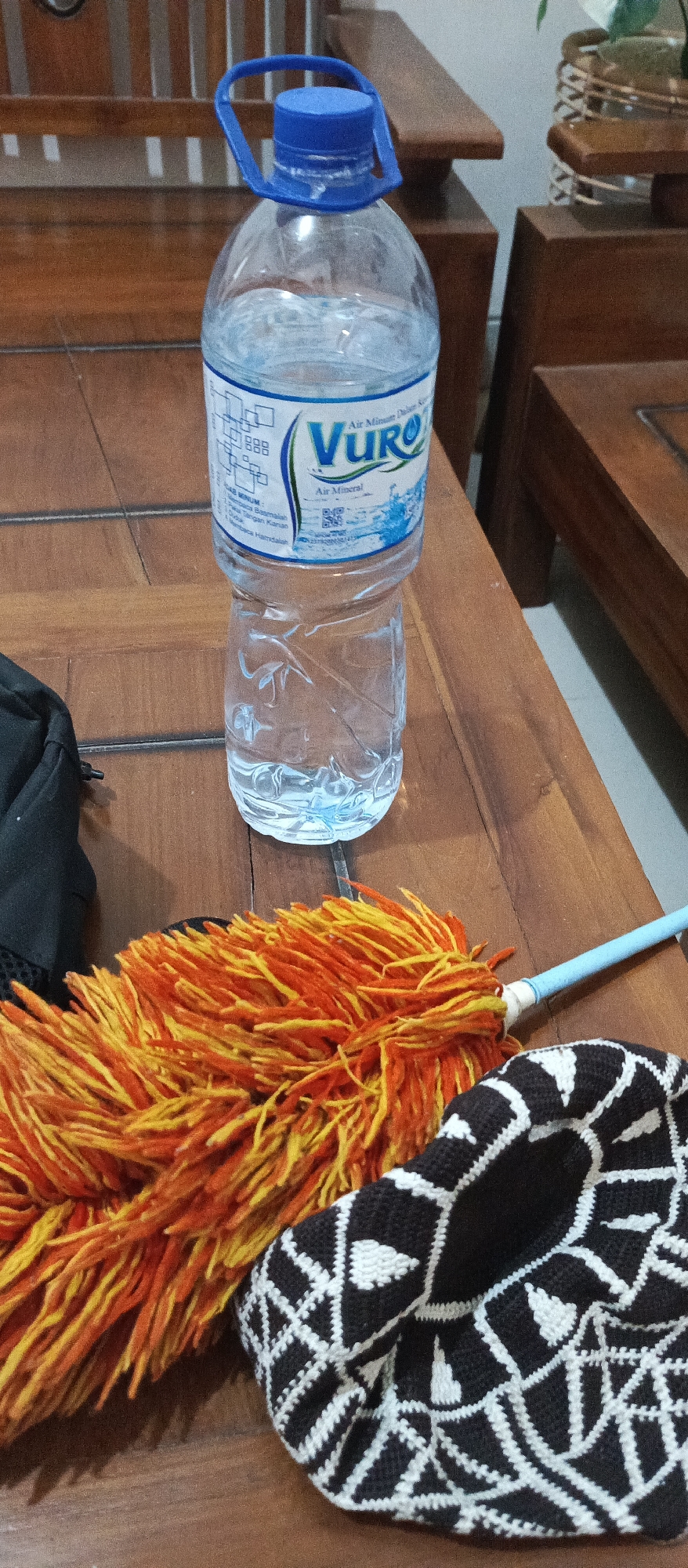 A low-angle, full shot shows a large bottle of VURO brand mineral water standing upright on a wooden surface, likely a table, in Jatisari, Indonesia. The bottle is partially filled with clear water and has a blue plastic handle and cap. A label on the bottle features the VURO logo and text in Indonesian. In the foreground, a fluffy duster with orange and yellow bristles is laid out, with its light blue plastic handle extending to the right. Partially beneath the duster is a black fabric item with a white geometric pattern. The background consists of a wooden chair and other wooden furniture. The lighting is dim, suggesting an indoor setting. There are no people visible in the image.