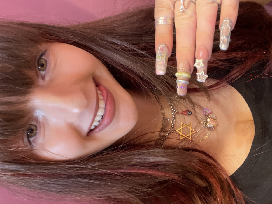 A close-up photograph features a smiling female subject looking directly at the camera. She has long brown hair with bangs and reddish highlights. Her lips are parted, showing teeth and a portion of her tongue. She is wearing a black top. Multiple layered necklaces are visible around her neck, including a silver-colored chain with letters, a prominent gold-colored Star of David pendant, a small red pendant with a blue eye motif, and other small charms including a heart. The subject's left hand is visible on the right side of the frame, displaying long, manicured fingernails with elaborate clear acrylic designs. Her fingers are adorned with numerous rings: a silver-toned band on the thumb, a silver-toned and a light green ring on the index finger, a large star-shaped ring with embedded rhinestones on the ring finger, and a textured silver-toned ring on the pinky finger. The background consists of a plain pink wall. The scene is located in the city of Raanana, Israel.