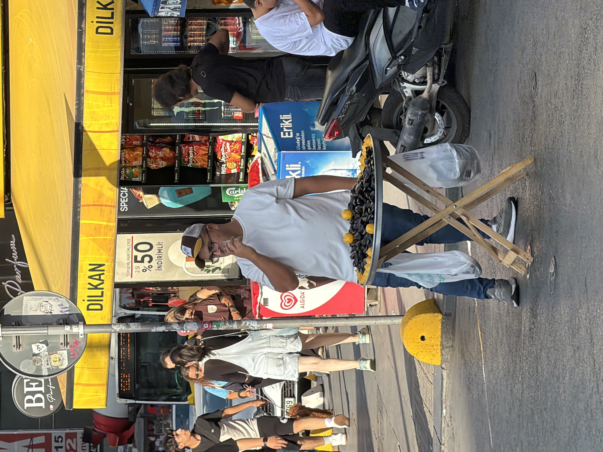 In Kadıköy, Turkey, on a bright, sunny day, a man stands by a sidewalk food stand, holding a cigarette to his mouth and looking down. He is wearing a white t-shirt, blue jeans, and grey sneakers. In front of him, a round tray sits on a wooden folding table, laden with mussels and garnished with yellow lemons. A white plastic bag hangs from the table leg. The background shows a busy street scene with people walking, a bus, a motorcycle, and a store with a yellow awning. The store has a sign that reads "DILKAN" and displays various food items and drinks. The overall impression is of a lively urban environment.