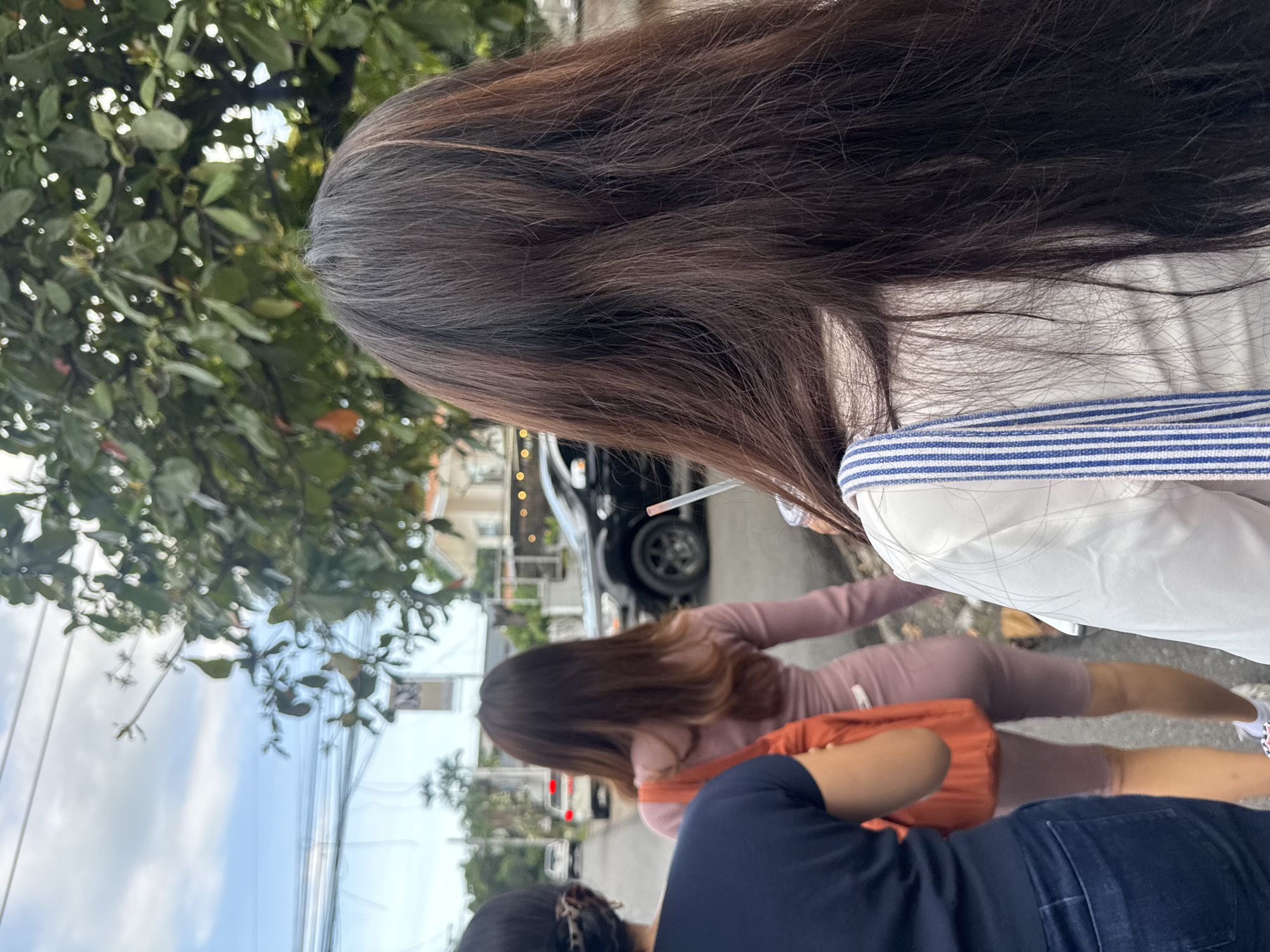 The image shows three women walking along a street. One is wearing a white top with a blue and white striped strap, and another is wearing a pink outfit. The third woman is wearing a dark blue shirt and jeans. There is a tree on the left side of the image, and a car is visible in the background. Based on visual features of street signs and architecture, the image may be located in Davao City, Philippines. One person appears to be smoking a cigarette or vaping.