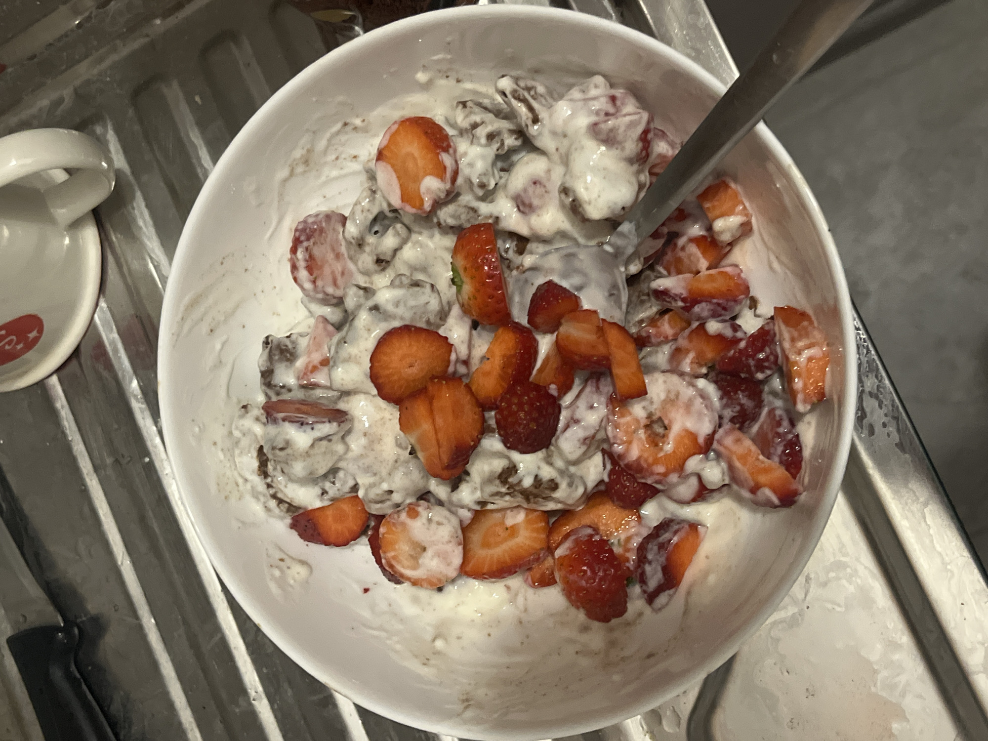 A white bowl containing a mixture of sliced strawberries, what appears to be chocolate pieces, and a creamy white substance (possibly yogurt or ice cream) sits in a metal sink. A metal spoon is partially submerged in the mixture. A white mug with a red design is visible to the left of the bowl. The setting appears to be a kitchen, with the sink made of stainless steel. There are no indications of the location being in Sofia, Bulgaria.