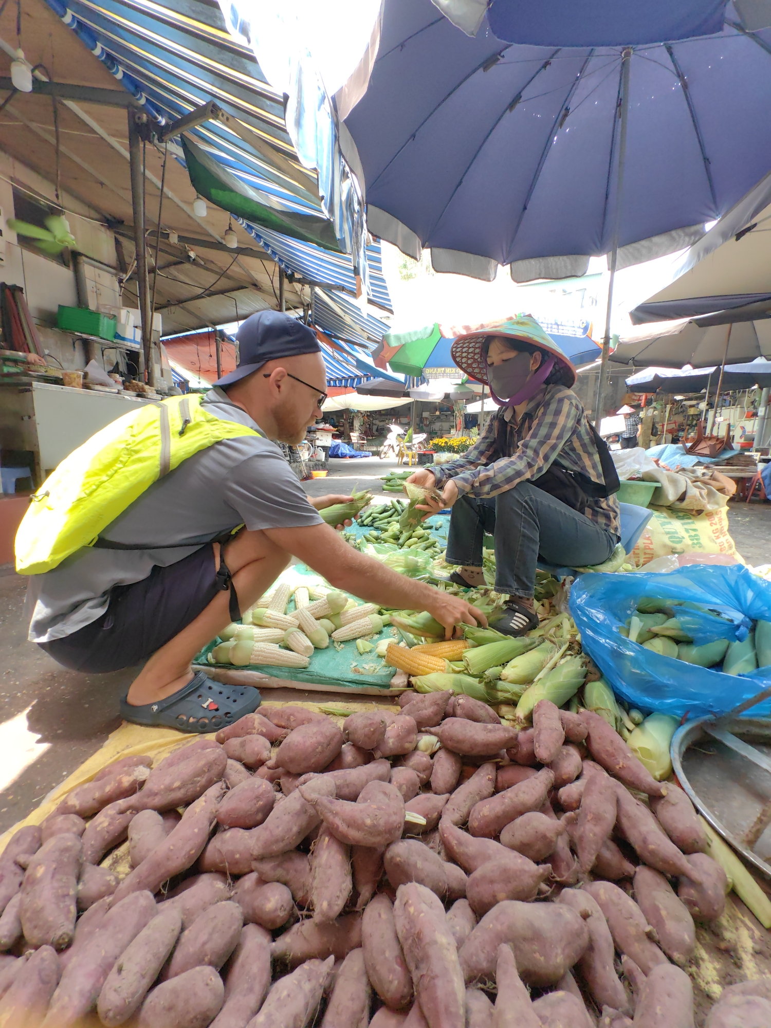 In Ha Long Ward, Vietnam, a man crouches at an outdoor market stall. The man, wearing a gray t-shirt, blue shorts, Crocs, and a blue baseball cap, has a yellow backpack. He is inspecting the corn on the cob at the stall. A woman wearing a conical hat, a plaid shirt, and a face mask sits on a bag behind a table loaded with corn and sweet potatoes. A blue plastic bag filled with produce sits to her right. A large pile of sweet potatoes is in the foreground. Umbrellas shade the market.