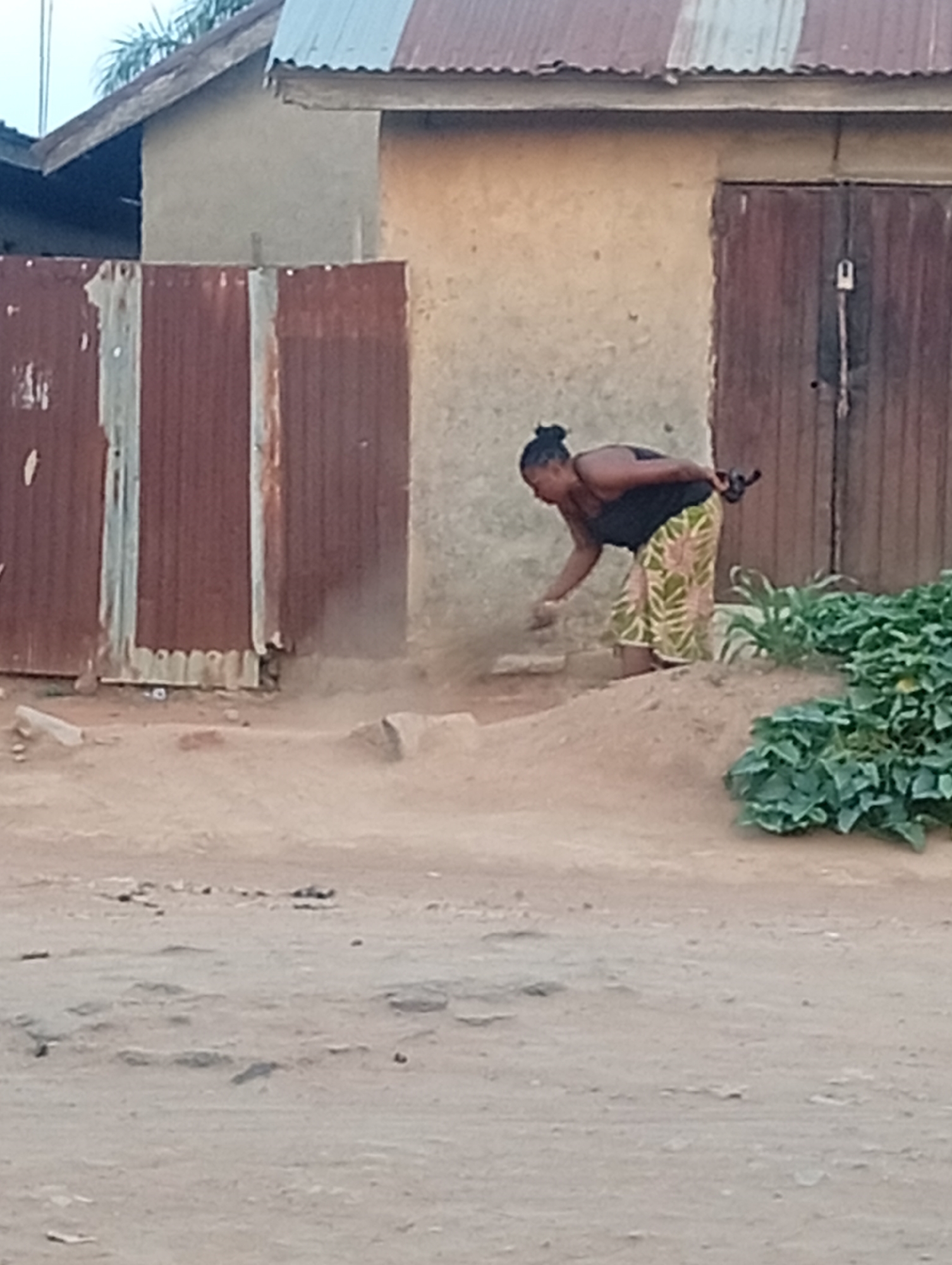 A woman is stooping to pick up something from the ground in front of a building with a rusted metal fence and a double door. The building appears to be located in a rural or undeveloped area, judging by the dirt ground and the lack of pavement. The city is Unknown, Unknown.