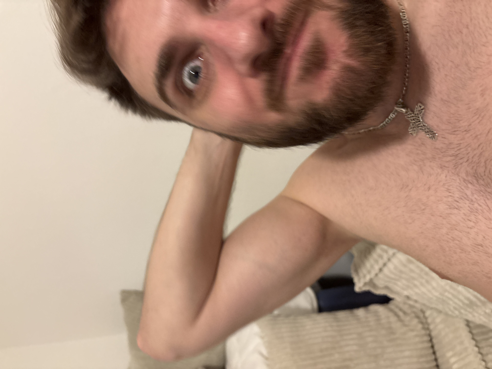 The image is a close-up, angled selfie of a male individual taken indoors. The man's face, shown from an upward and slightly rotated perspective, occupies the left side of the frame. He has light skin, short dark brown hair, and a beard or heavy stubble. His left eye, a clear light blue, is wide open and looks directly into the camera. Around his neck, a silver-colored chain with a detailed, possibly ornate, cross pendant is visible, resting against his chest.
The man appears to be shirtless, with his right shoulder and part of his upper arm visible on the right side of the image. His left arm is bent, with his hand likely supporting his head, though only the arm is clearly depicted.
The background consists of plain, light-colored walls, likely off-white or cream. In the lower right, a neutral-toned, textured fabric, possibly a blanket or bedding with a ribbed pattern, is visible, suggesting an indoor setting like a bedroom. The lighting is soft and ambient. The image has a slight overall blur, potentially due to motion or shallow depth of field. The man's expression is neutral and direct. There is no visible text in the image. No specific details within the image indicate its location as Mönchengladbach, Germany.