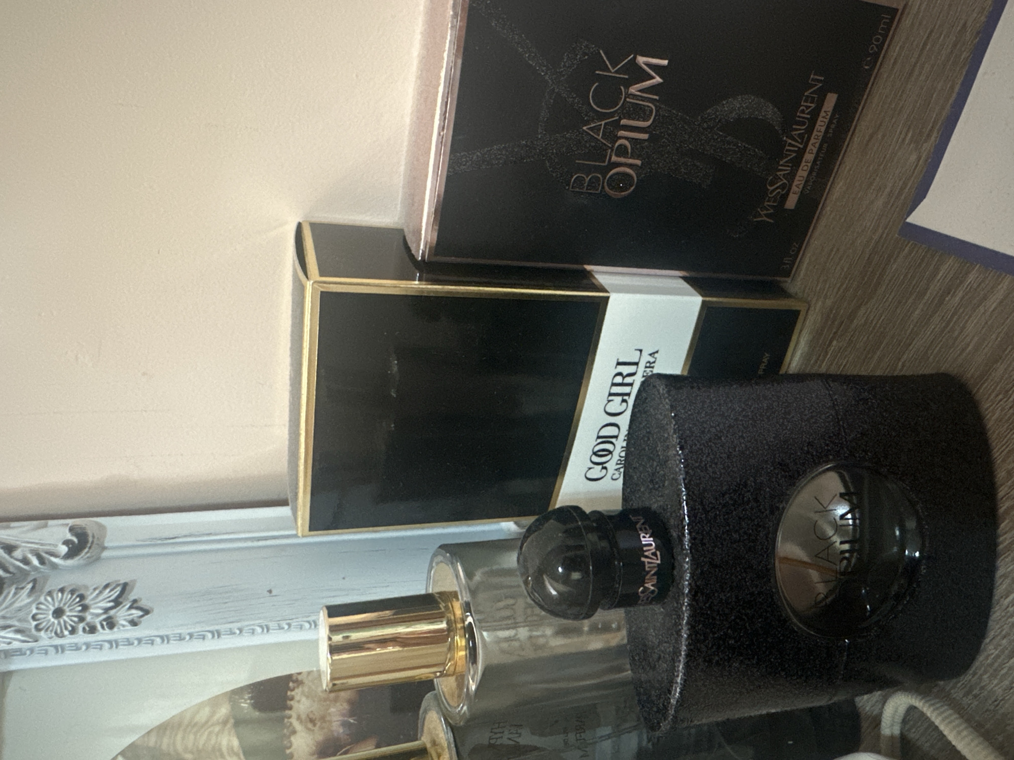 A still life photograph depicts a collection of perfume bottles and their boxes. At the top is a black box labeled "BLACK OPIUM YVES SAINT LAURENT EAU DE PARFUM VAPORISATEUR SPRAY 90ml" At the middle, there is a black box labeled "GOOD GIRL CAROLINA HERA" At the bottom, there is a perfume bottle labeled "SAINT LAURENT". The objects rest on a surface with the bottom left corner showing a white decorative frame. The photograph appears to be taken indoors under artificial lighting. The location and city details are not discernible from the image.