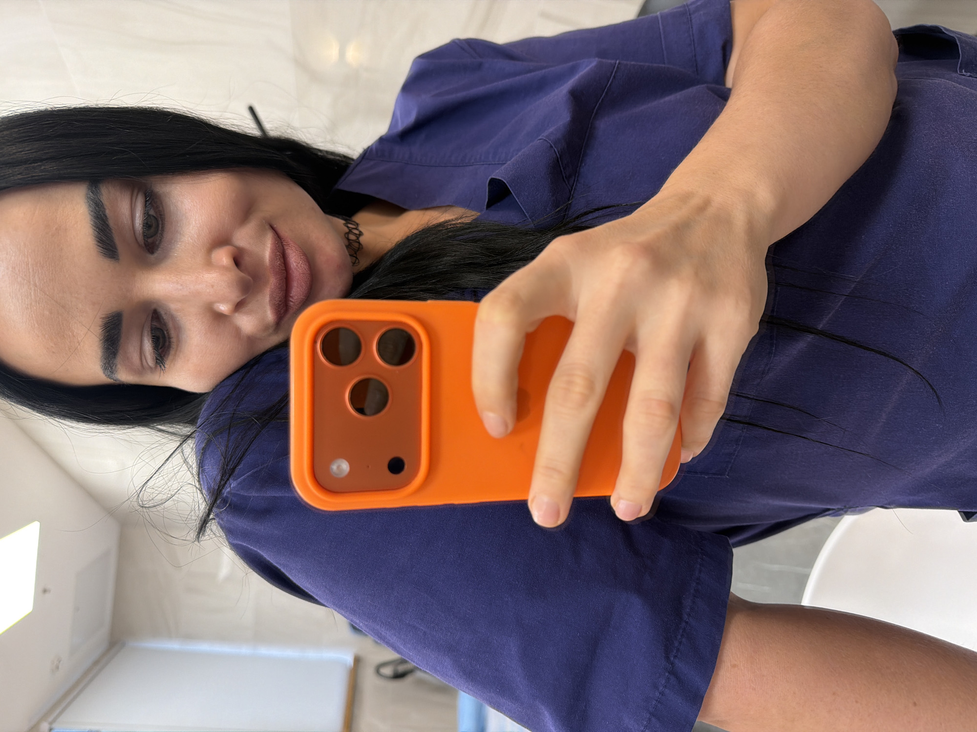 The image, presented rotated counter-clockwise, depicts a woman taking a selfie in an indoor setting, likely in Saint Petersburg, Russia. Described as if upright, the primary subject is a woman with long, straight dark hair, neatly defined dark eyebrows, and brown eyes, looking towards her phone with a subtle, closed-mouth smile. She is wearing a dark blue V-neck scrub-style top, suggesting a professional or medical environment, and a thin black choker necklace.
She holds an orange-cased smartphone in her right hand, positioned to capture her reflection or a selfie. The back of the phone case, featuring a prominent camera module with three lenses, is clearly visible. Her fingers are gently wrapped around the device. The background is simple and well-lit, composed of light-colored, possibly white, walls and subtle architectural details, indicating a clean and modern indoor space. The bright, even lighting suggests daytime. No discernible text is visible in the image.