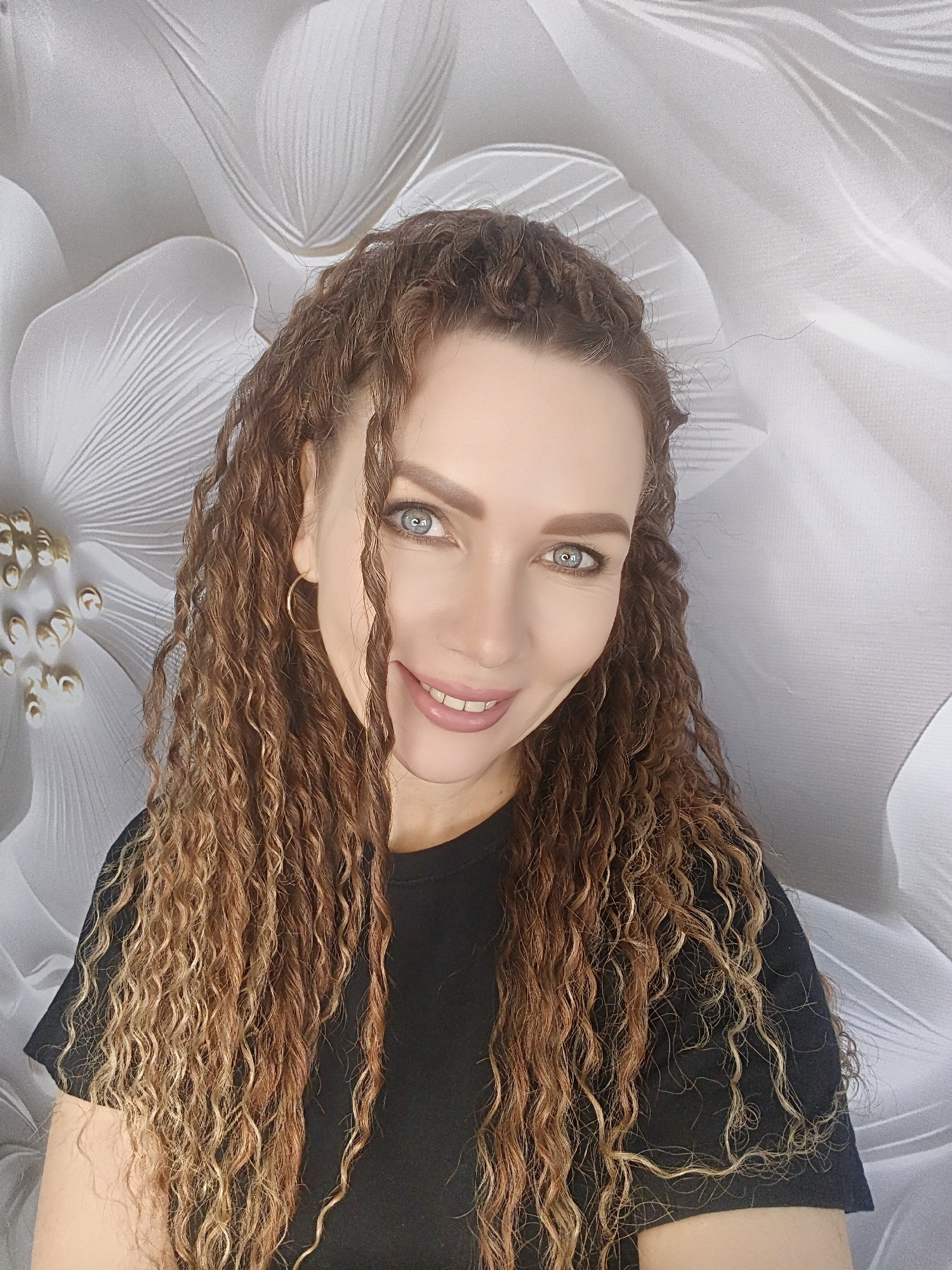 A woman with long, curly brown hair and blue eyes smiles at the camera. She wears a black shirt and a gold hoop earring is visible in her left ear. The background features a large, white floral design. The location is Unknown, Unknown.